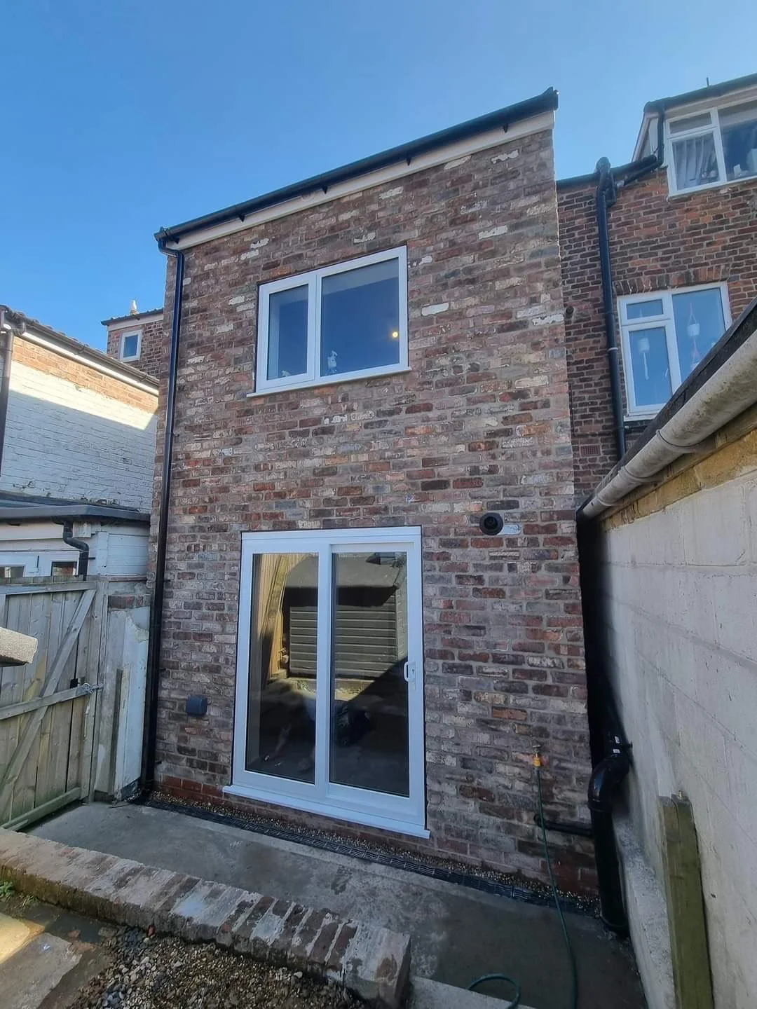 Back of a brick house with two large white-framed windows and a white sliding glass door. A small garden area and a wooden fence are visible in the foreground.