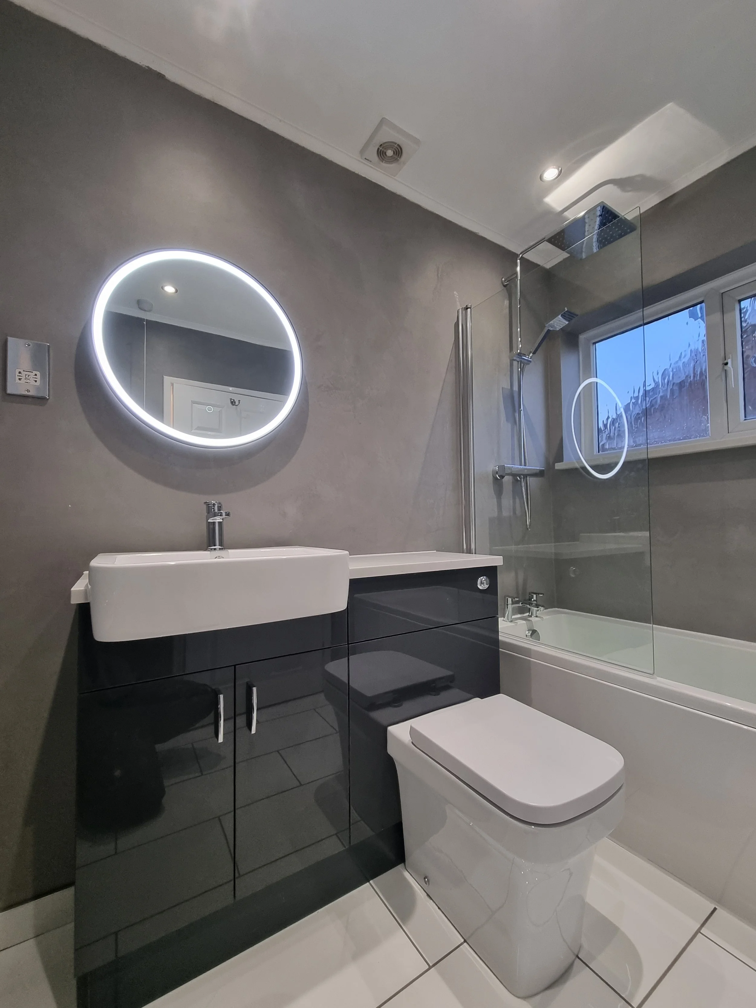 Modern bathroom with a round illuminated mirror, a white sink, a dark vanity, a toilet, a bathtub with a glass shower screen, and a window, all in a gray and white color scheme.