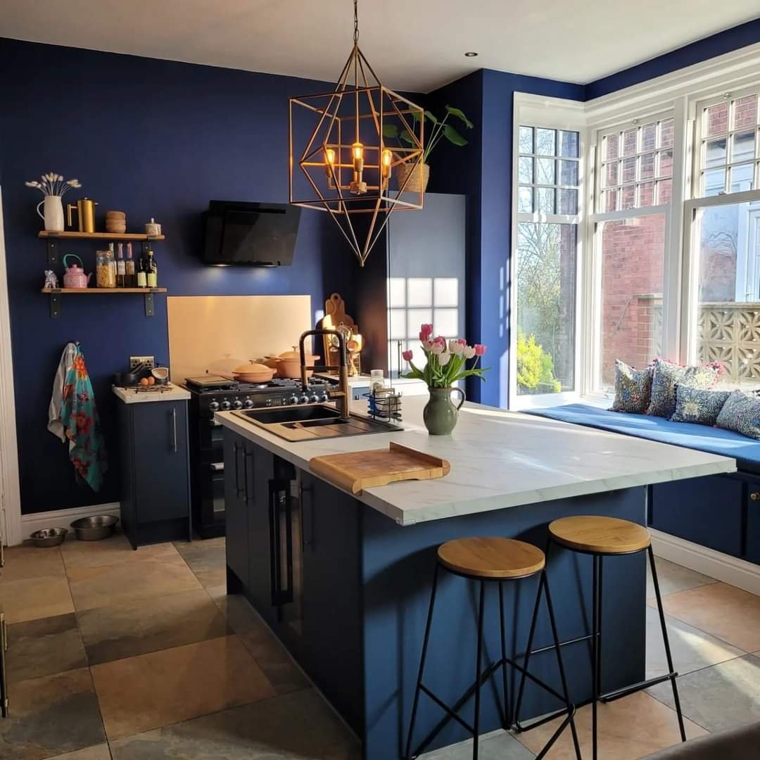 A modern kitchen with dark blue walls, a white marble island, and large windows. There are two wooden bar stools, a black faucet, and a vase of pink flowers on the island. Floating shelves hold jars and decorative items, and a small TV is mounted on 