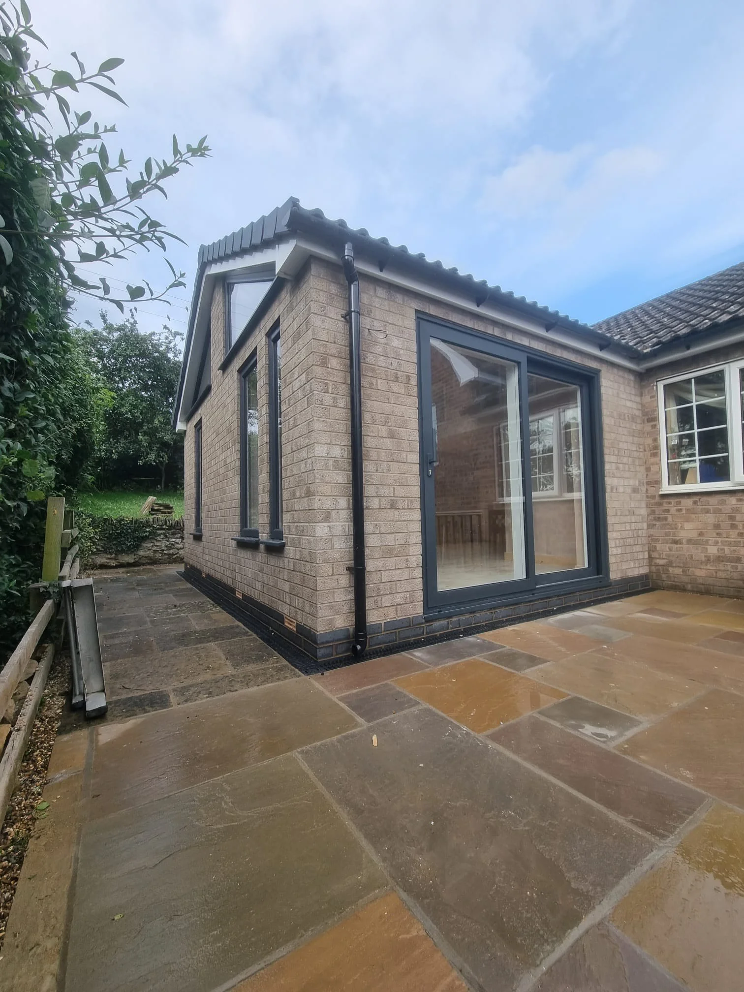 Exterior view of a brick house with large sliding glass doors and multiple windows, paved patio, and a cloudy sky.