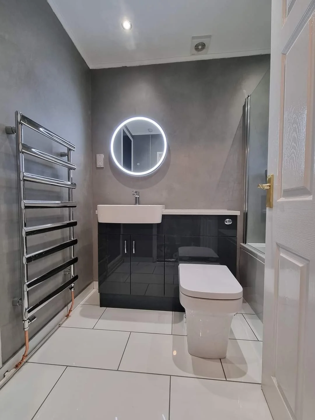 Modern bathroom with a heated towel rail, a circular backlit mirror, a rectangular sink with a black vanity, and a toilet next to a glass shower enclosure, with white tiled flooring and gray walls.