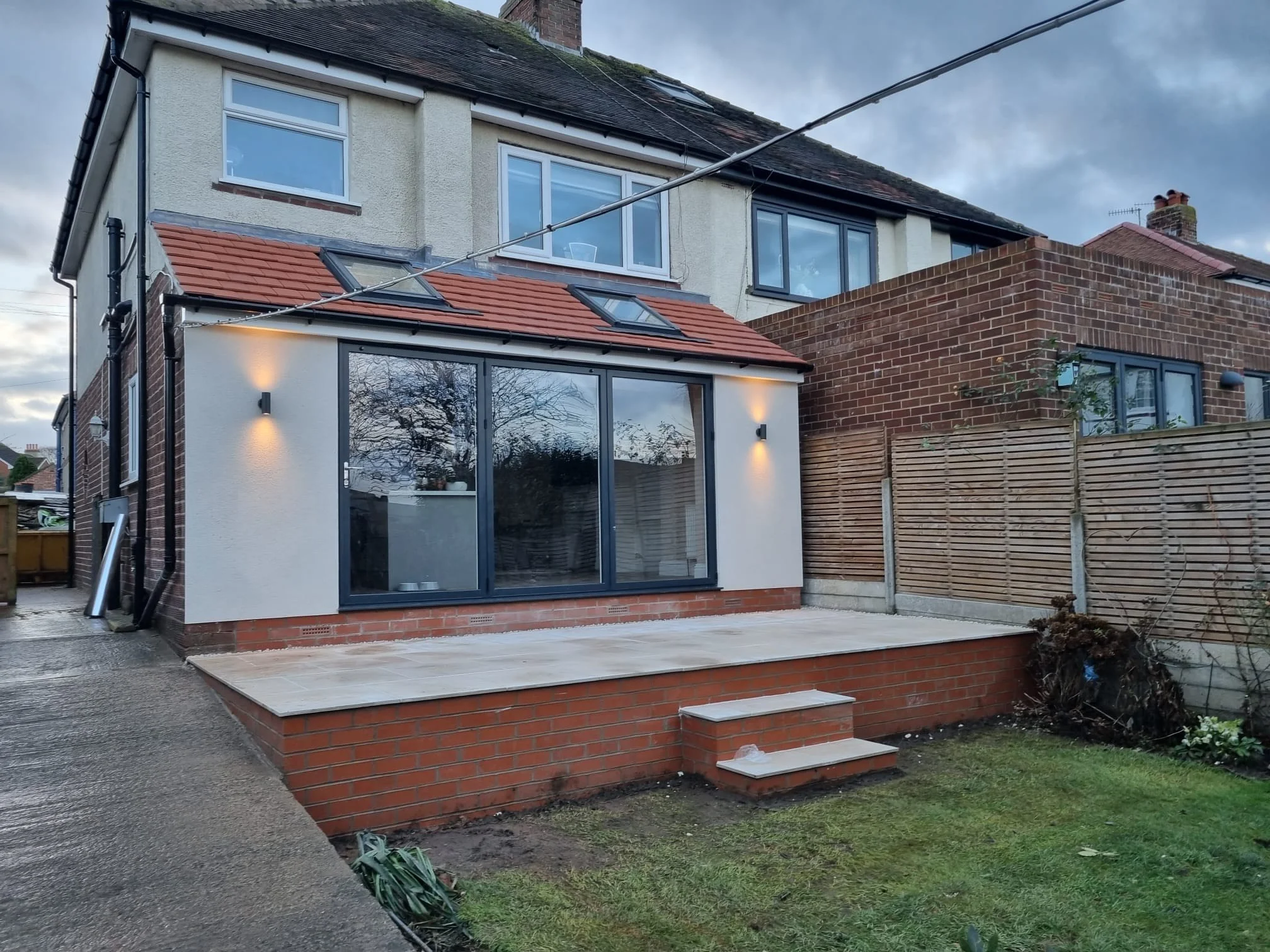 Backyard of a house with a newly built patio area, large sliding glass doors, outdoor wall lights, a small set of steps, and a lawn with some bushes on the side.