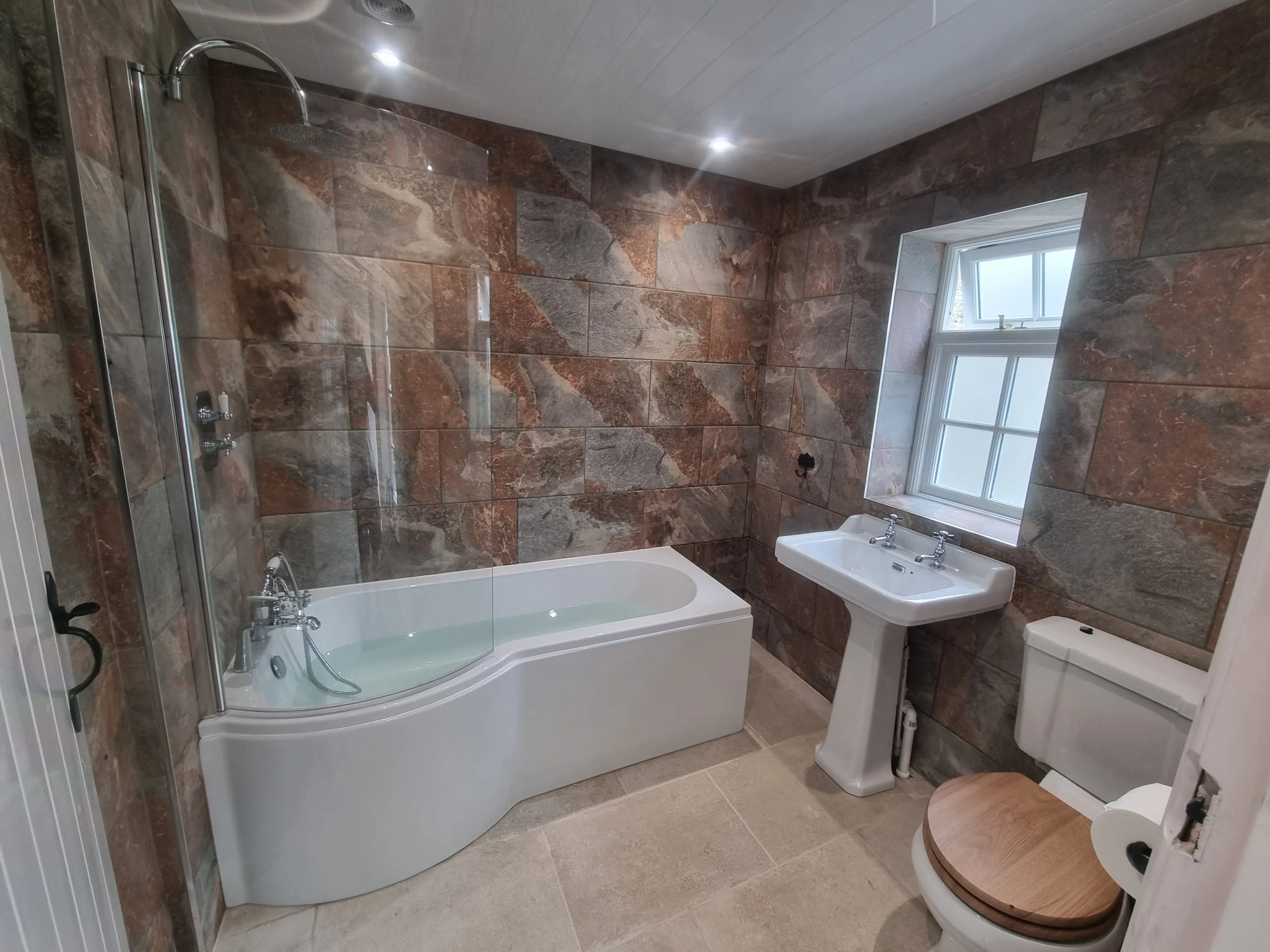 A bathroom with brown and gray stone walls, a white bathtub with a curved front, a glass shower screen, a white pedestal sink, a toilet with a wooden seat, and a small window with frosted glass. The floor is tiled in a light beige color.
