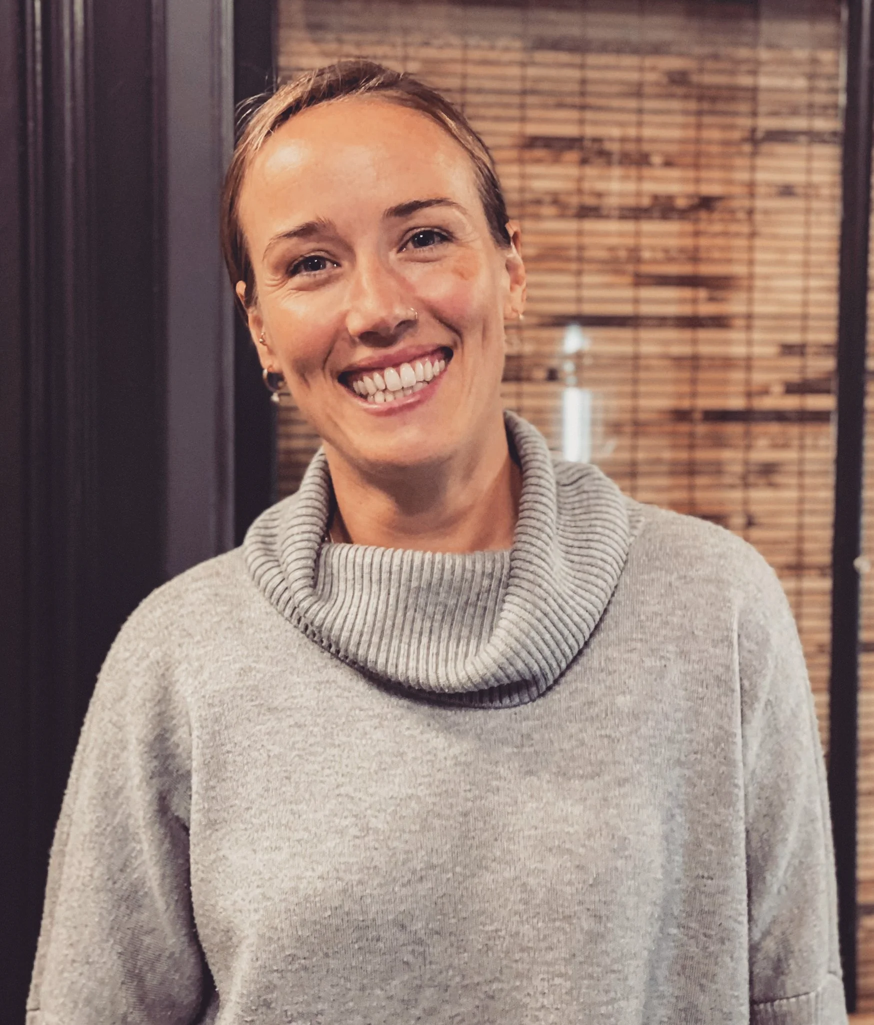 A woman with short hair and a gray turtleneck sweater, smiling indoors with wooden blinds in the background.