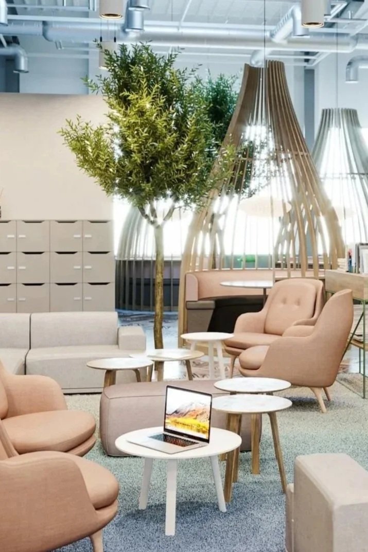 Modern office lounge area with comfortable pink and beige armchairs, small white side tables, a laptop, a large potted tree, and stylish wooden dividers forming semi-private booths, all under an industrial ceiling with visible ductwork.