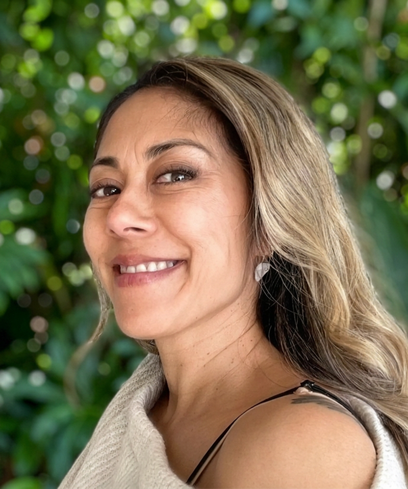 Close-up of a woman smiling outdoors with trees in the background.