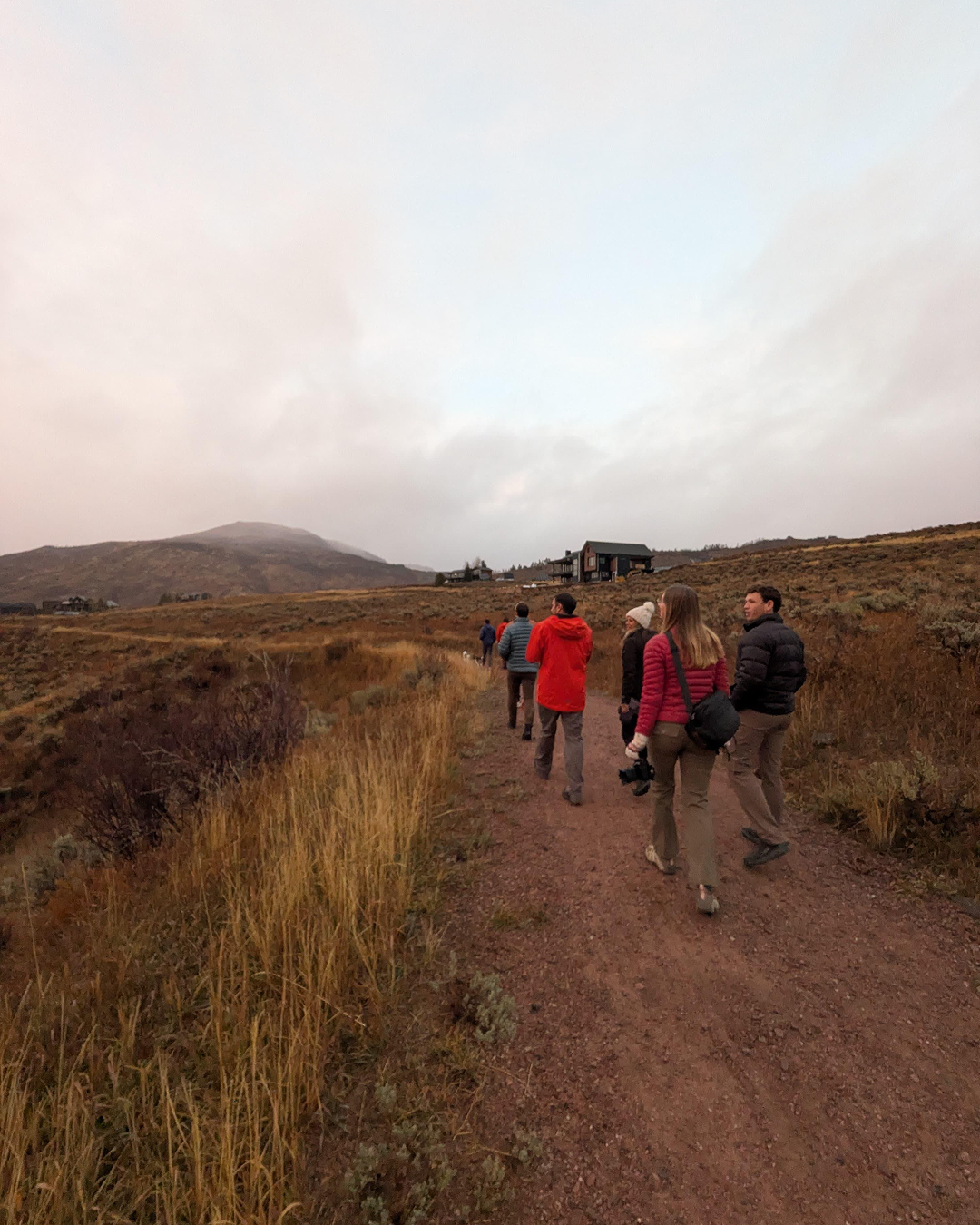 Group of people hiking on a dirt trail in a mountainous landscape with dry grass and a cloudy sky.