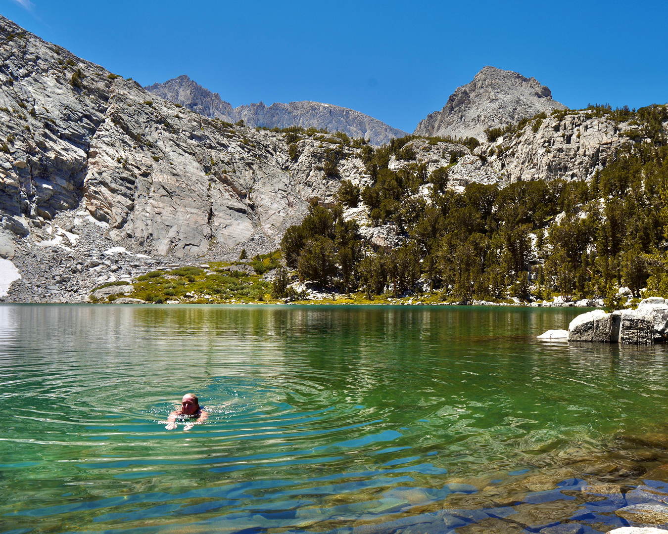 Person swimming in a mountain lake with rocky cliffs, green trees, and clear blue sky in the background.
