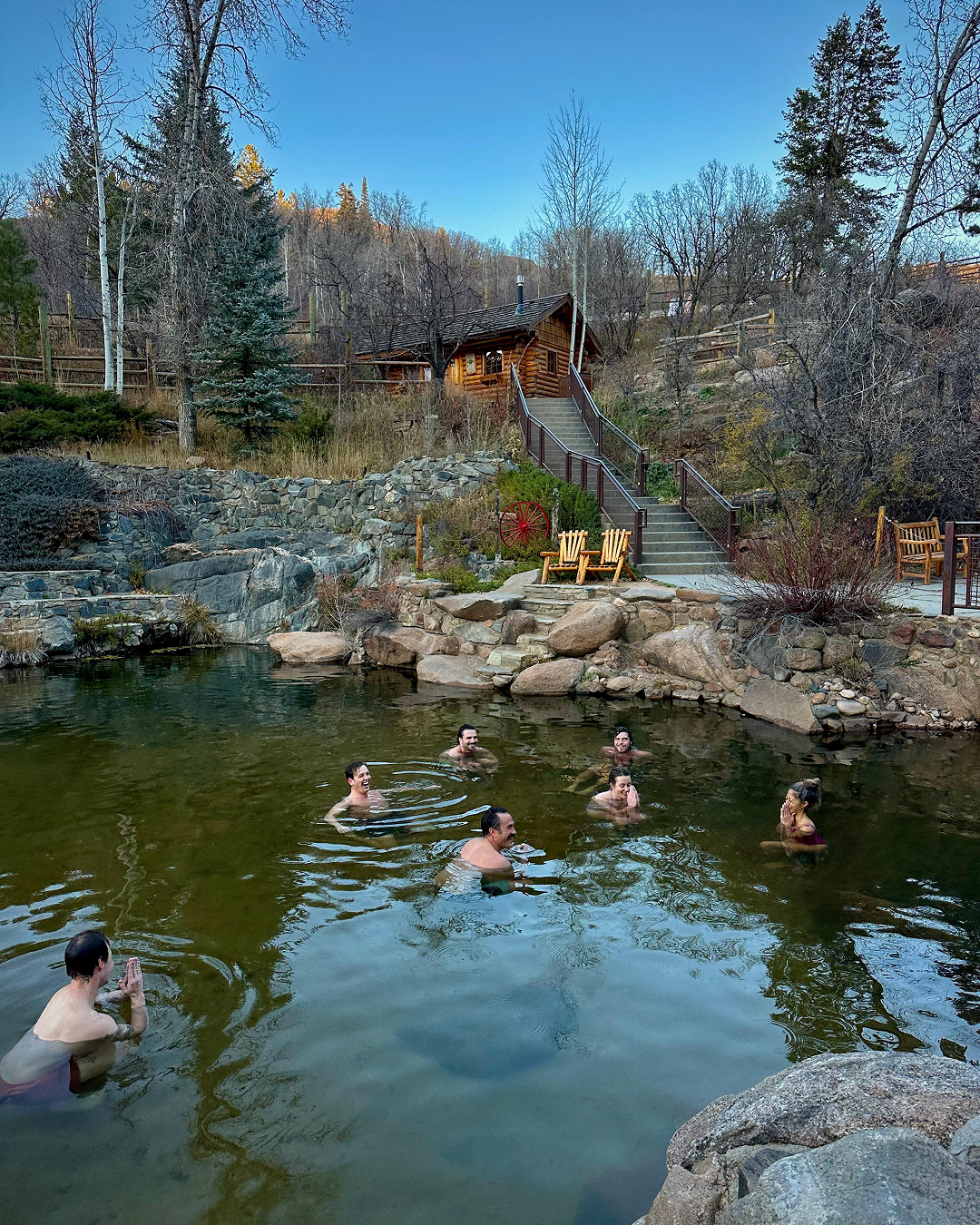 People enjoying a hot spring pool surrounded by rocks and nature, with a wooden cabin and stairs in the background.