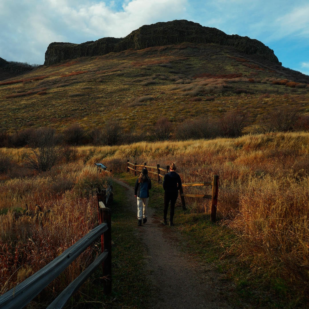 Two people walk on a dirt trail through a grassy hillside with a mountain in the background under a partly cloudy sky.