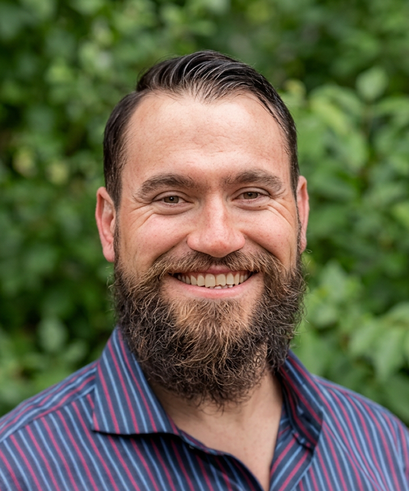 Portrait of a smiling man with a beard and short, slicked-back hair, wearing a striped blue and red shirt, outdoors with green foliage in the background.