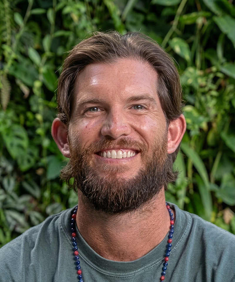A smiling man with a beard and brown hair, wearing a beaded necklace and a gray t-shirt, standing outdoors in front of green foliage.