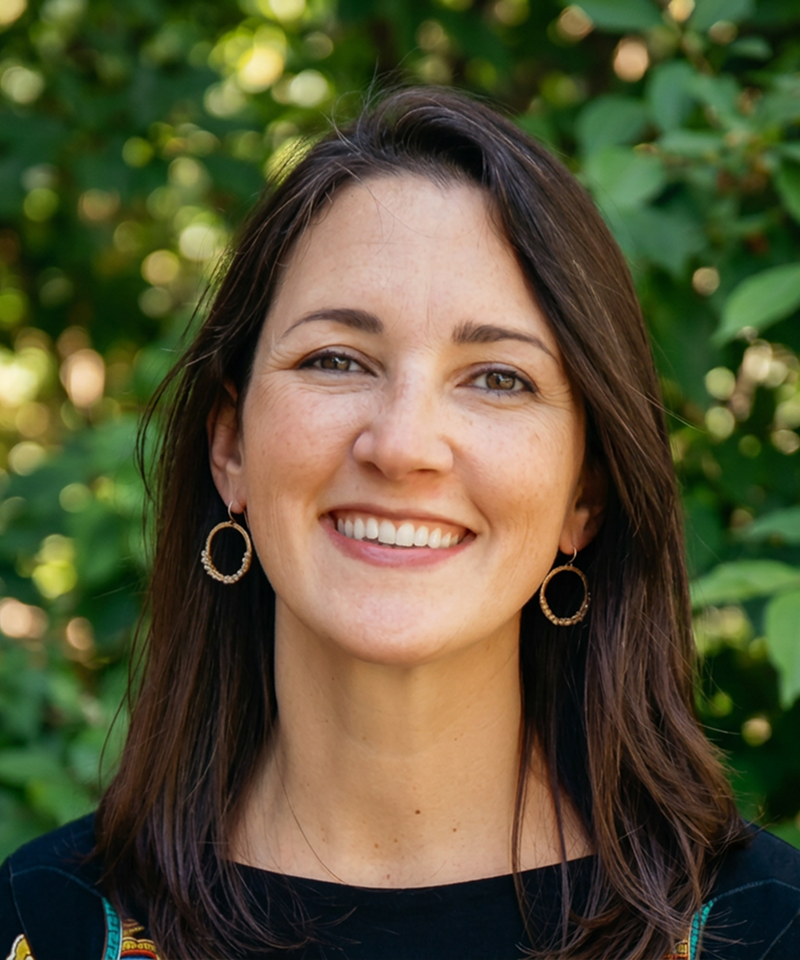 A woman with shoulder-length brown hair, wearing earrings and a black top, smiling outdoors with a background of green foliage.