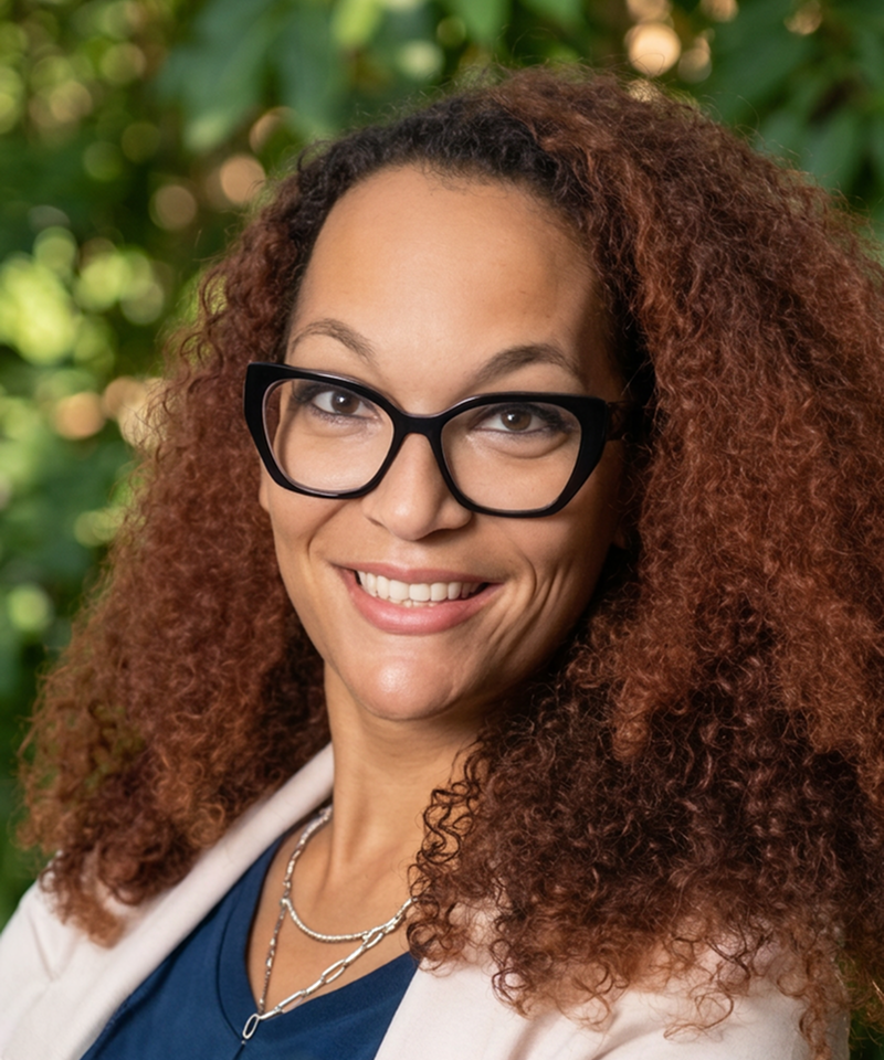 A smiling woman with curly brown hair, wearing glasses, a navy blue top, and a white blazer, outdoors with greenery in the background.