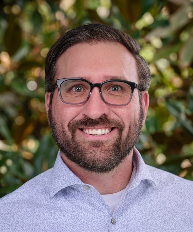 A man with brown hair and glasses smiling in front of green foliage.
