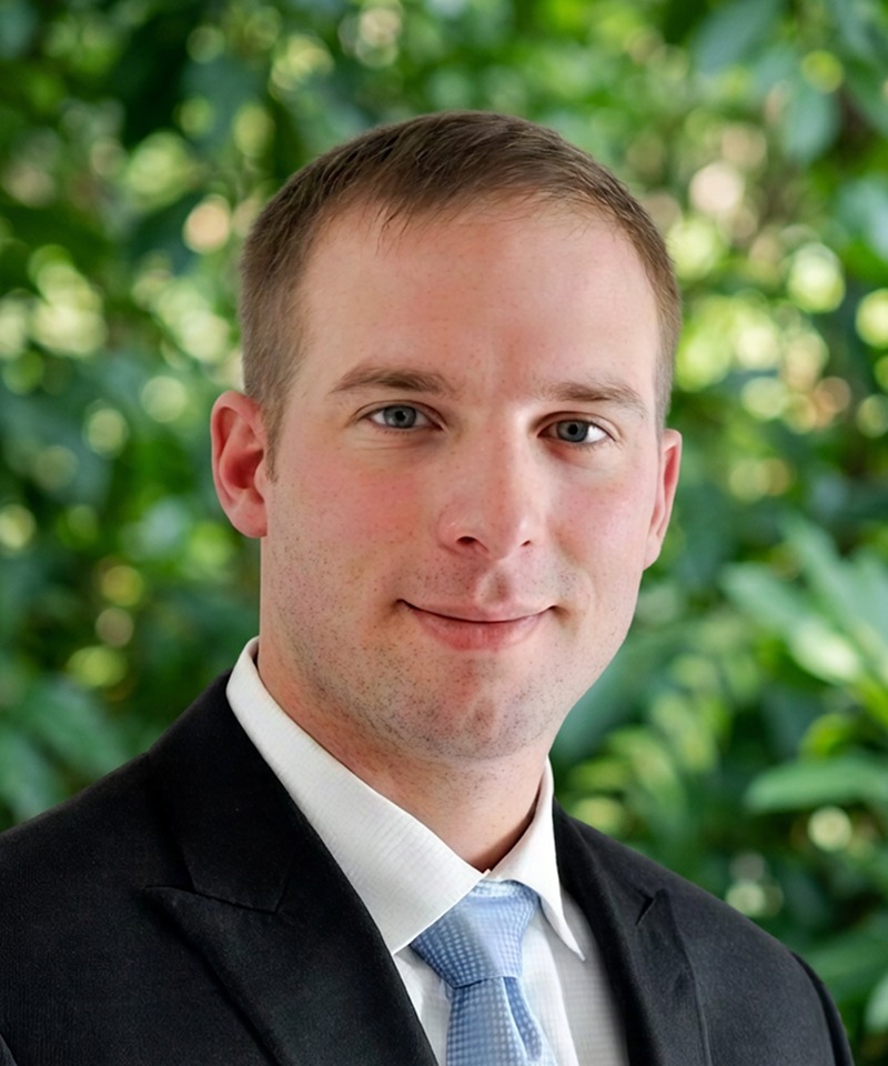 Headshot of a young man with short brown hair, blue eyes, and fair skin, wearing a black suit, white dress shirt, and light blue tie, against a blurred green leafy background.
