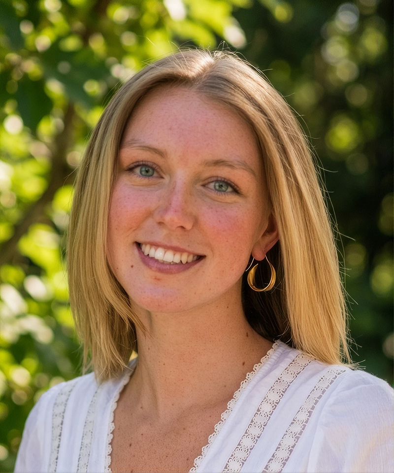 A young woman with blonde hair, light skin, blue eyes, and a bright smile, standing outdoors with greenery in the background.