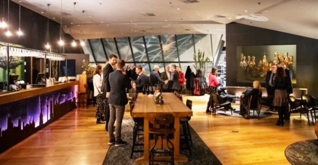 Mennesker som samles i en moderne, innendørs restaurant eller lounge med trebord, bar, og kunst på veggen.