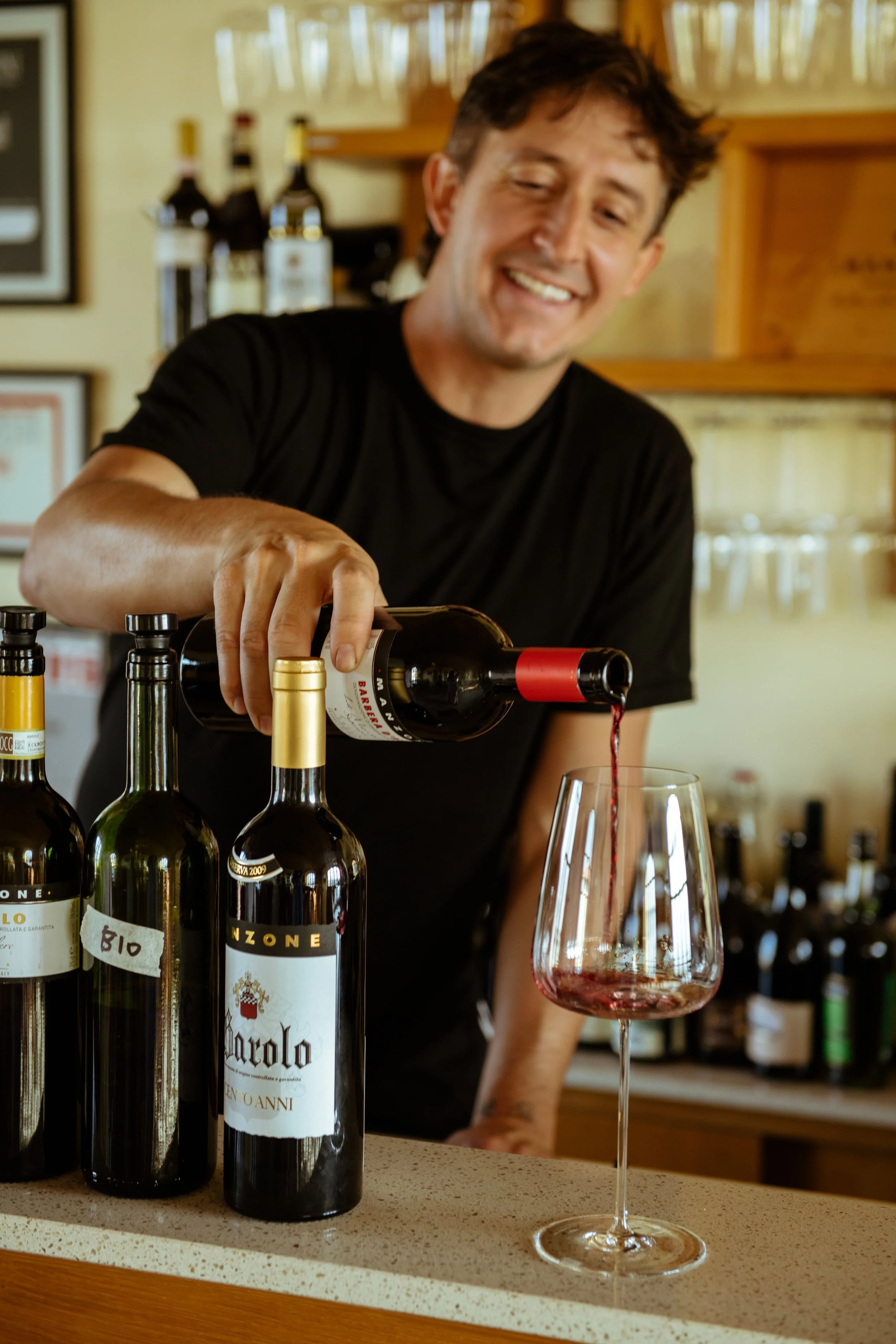 A man pouring red wine into a glass at a bar or restaurant with bottles of wine behind him.