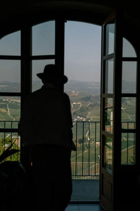 Silhouette of a person wearing a hat looking out through an open door at a landscape view of rolling hills and fields.