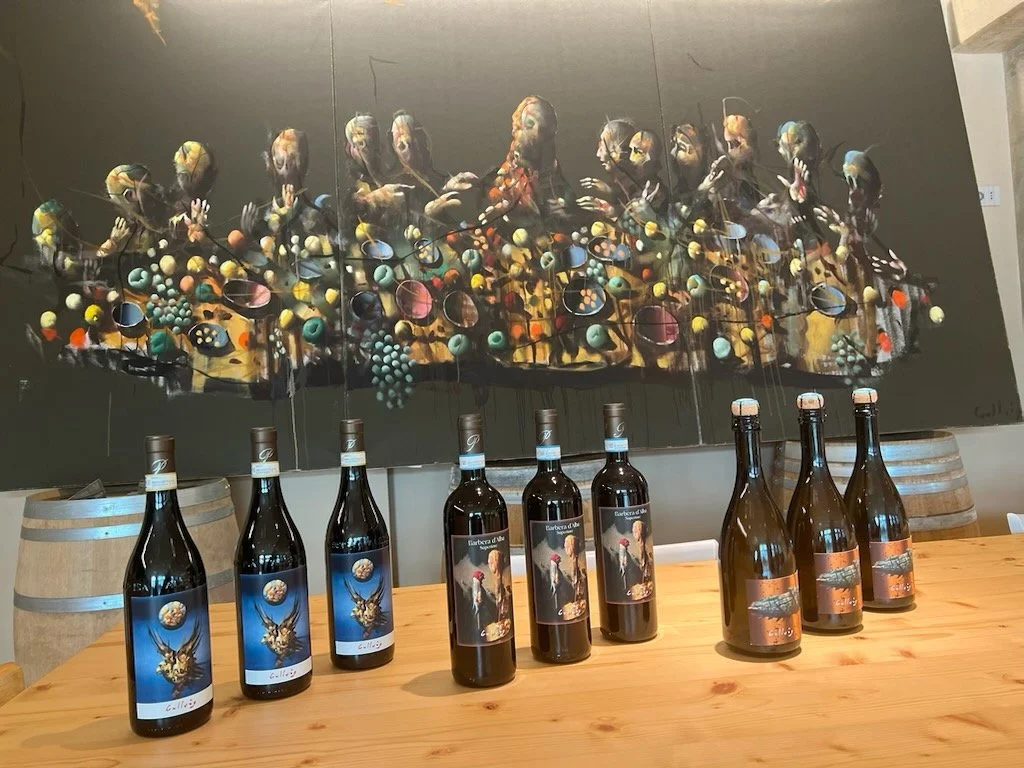A row of wine bottles with themed labels on a wooden table in front of a large abstract painting, which features multiple human faces and colorful, circular shapes, with barrels to the side. Håkon Gullvåg art.