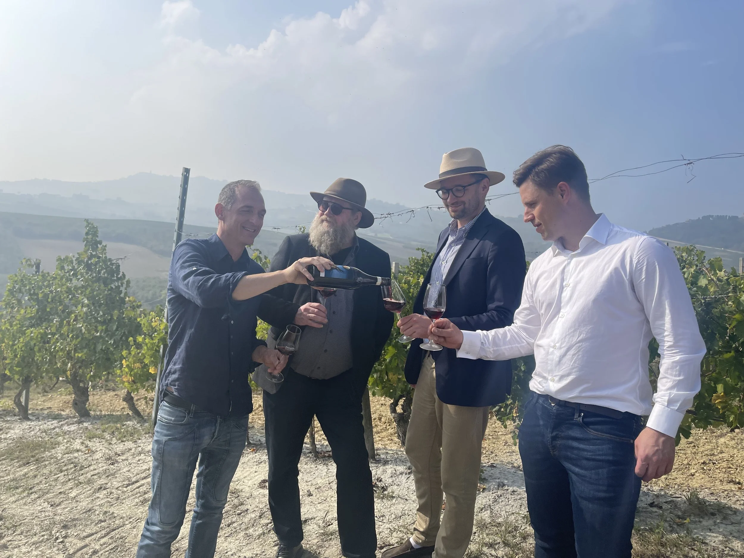 Fire menn som smaker og nyter vin i en vinmark om dagen.