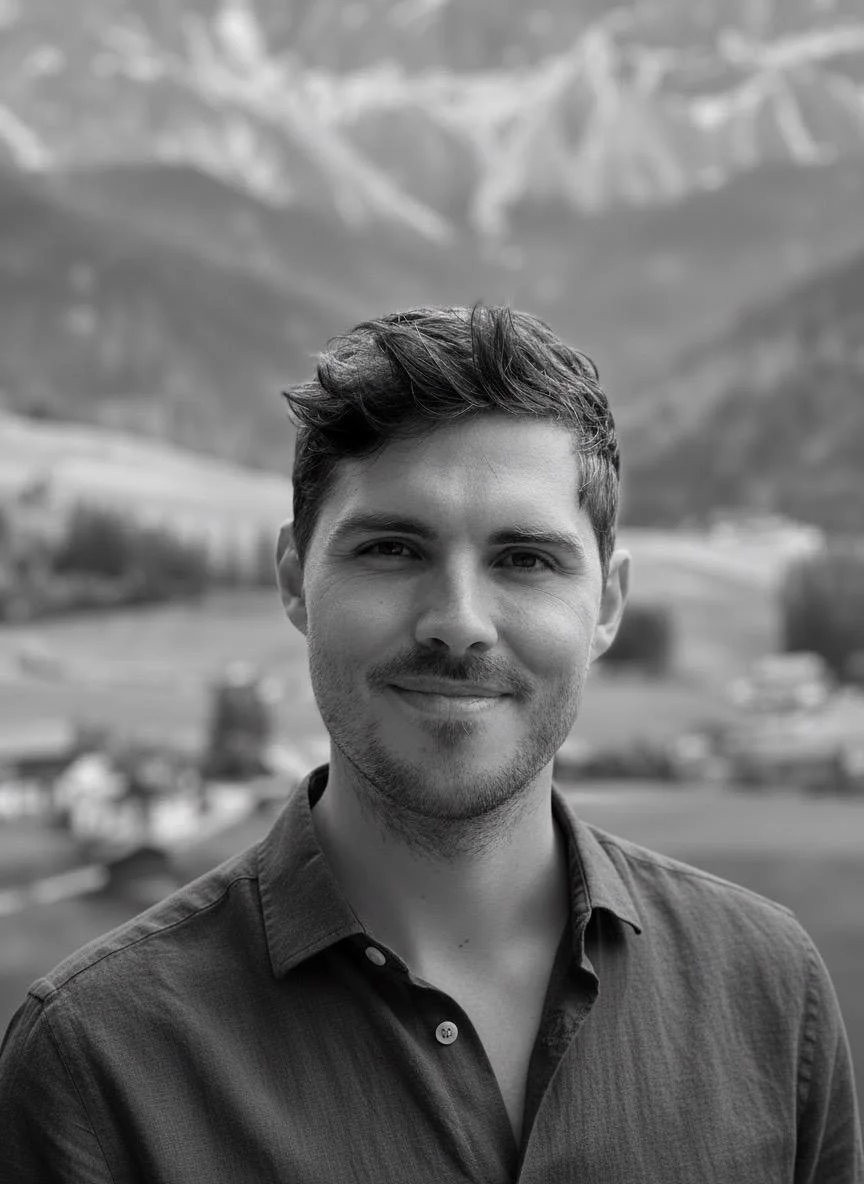 Ein junger Mann mit dunklem, leicht gewelltem Haar, trägt ein offenes Hemd und lächelt. Im Hintergrund sind Berge und ein Tal zu sehen.