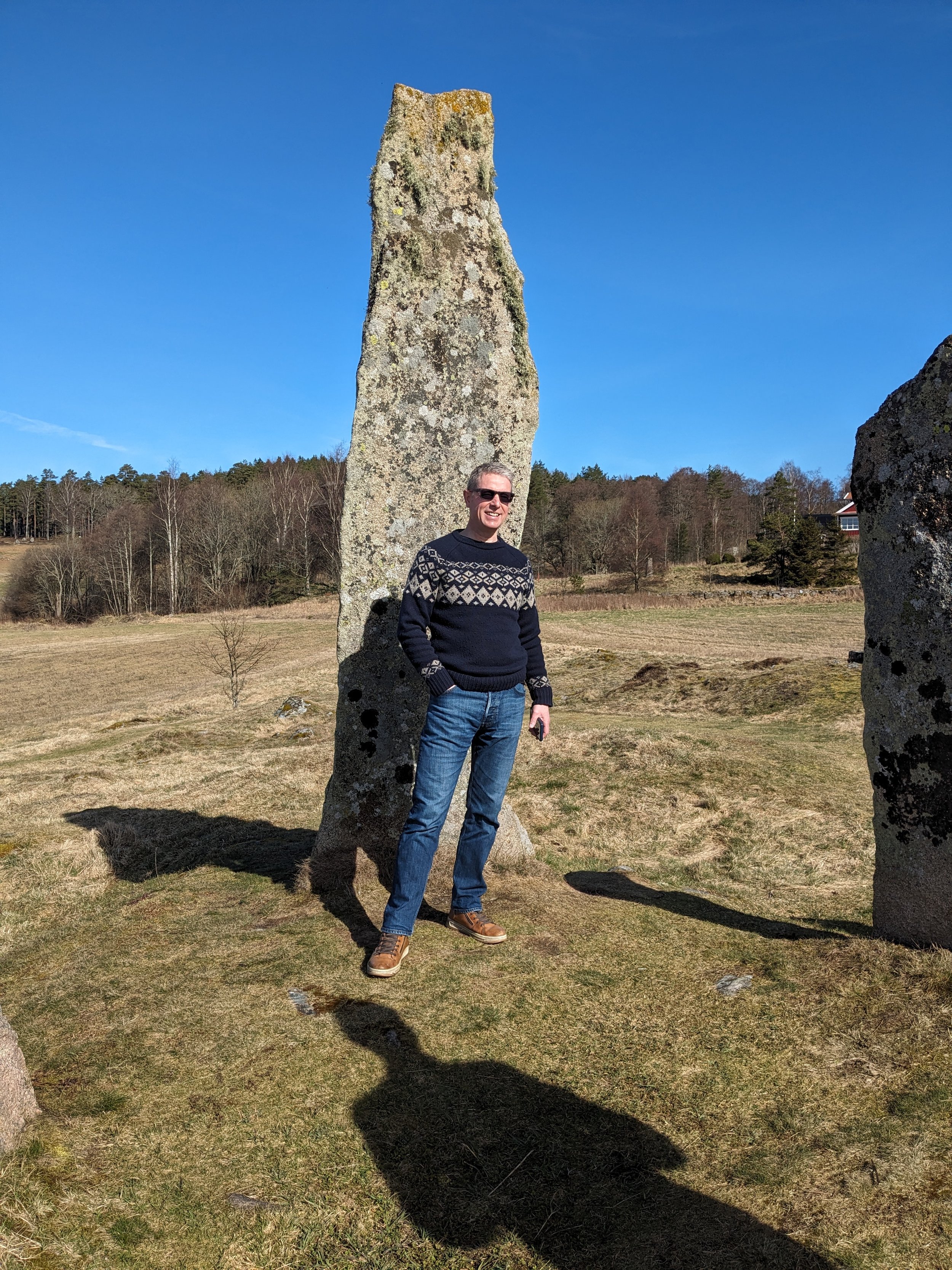 A man standing next to a tall, irregularly shaped standing stone outdoors on a grassy field with trees and a house in the background, under a clear blue sky.