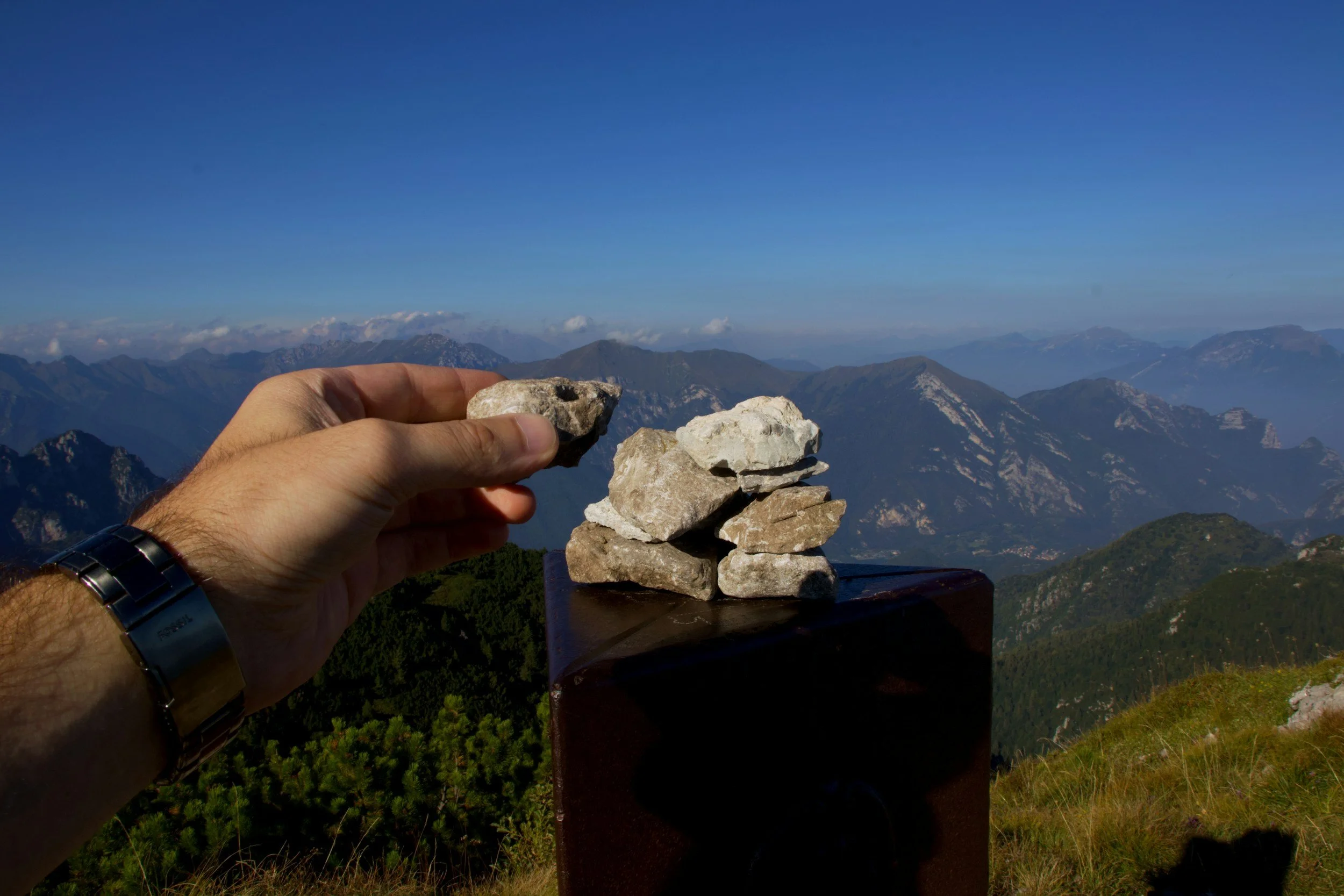 A person's hand stacking rocks on a wooden pedestal against a mountainous landscape with blue sky and distant clouds.