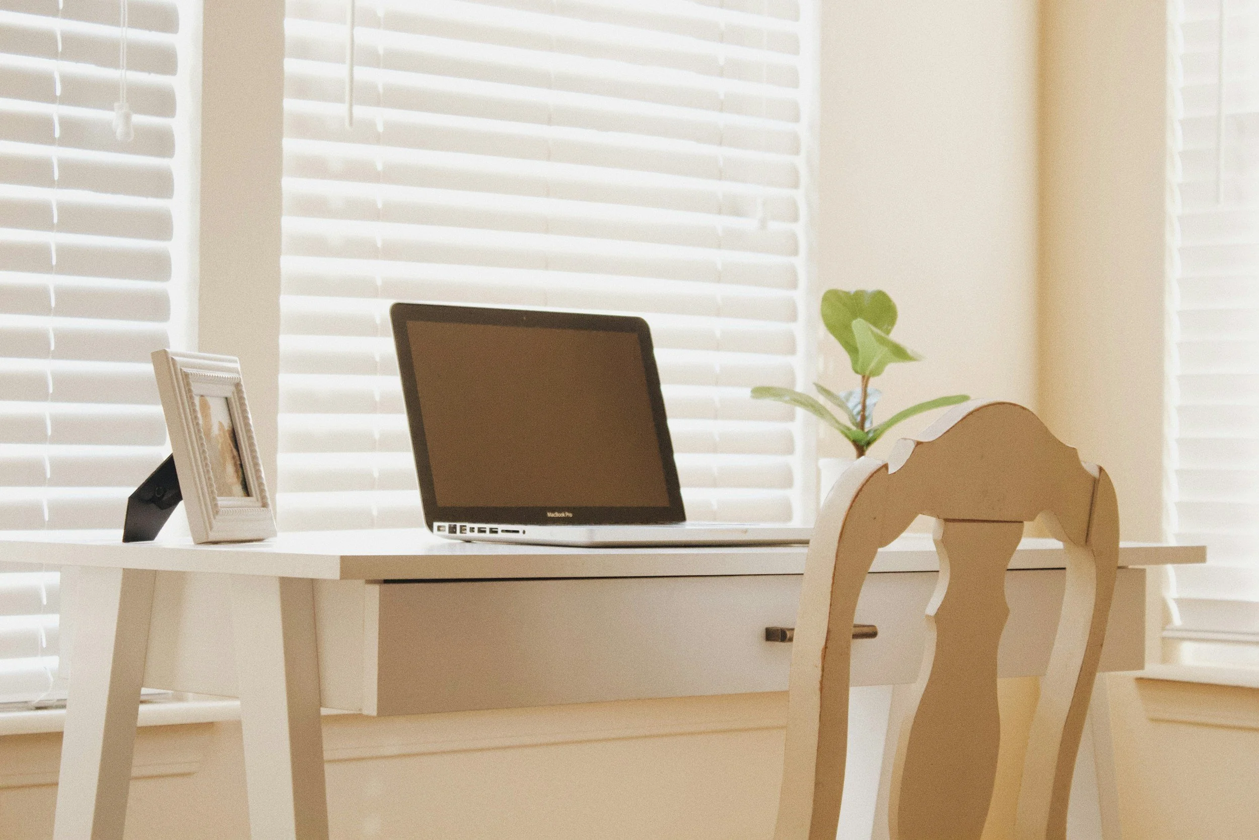 A white desk with a laptop, a picture frame, and a potted plant, in front of large window blinds in a bright room.