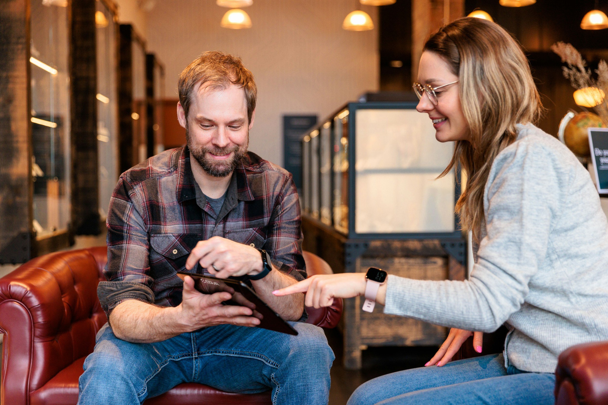 A man and a woman sitting on a leather couch in a cozy, modern cafe or coffee shop, smiling and looking at a tablet device together.