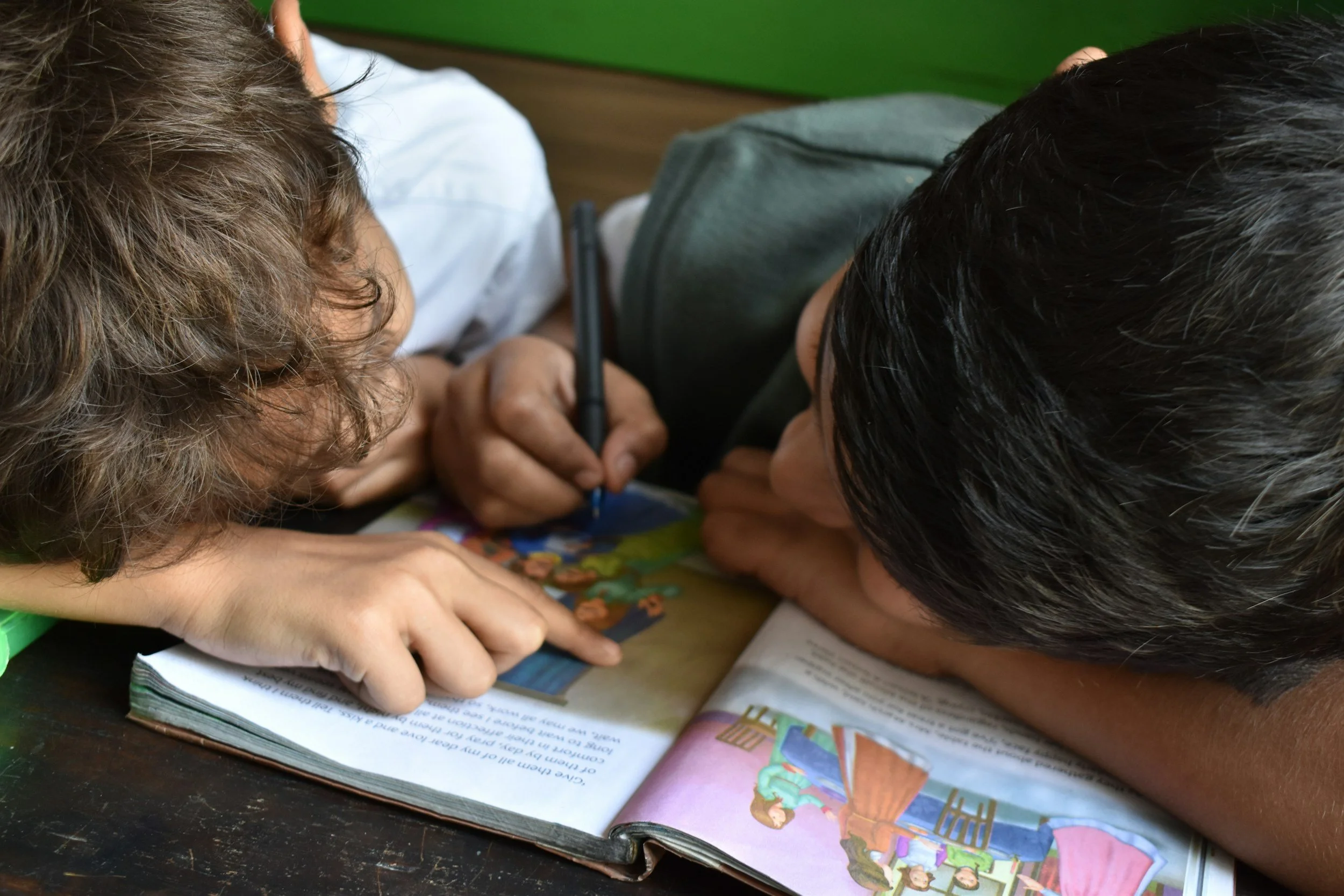 Two children lying on a table, closely looking at an open book with colorful illustrations and reading together.