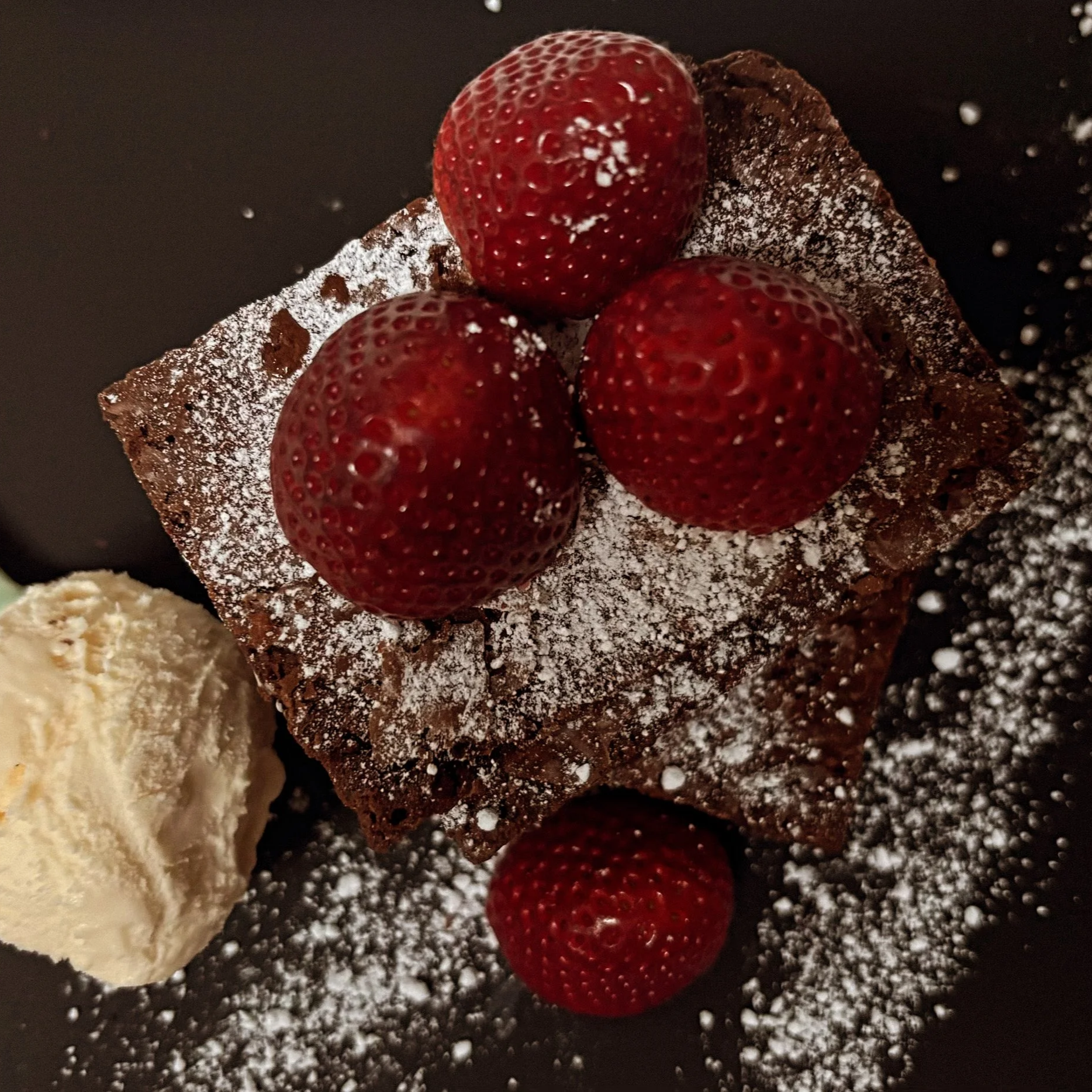 Chocolate waffle topped with three strawberries and powdered sugar, served with a scoop of vanilla ice cream on the side.