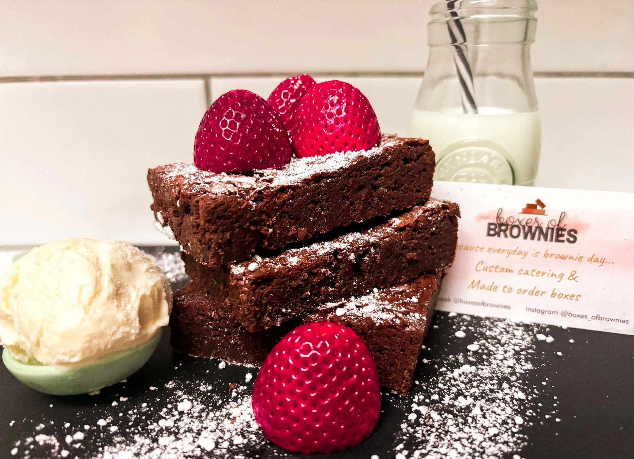 Three stacked chocolate brownies topped with fresh strawberries and powdered sugar, served with a scoop of vanilla ice cream and a bottle of milk in the background.