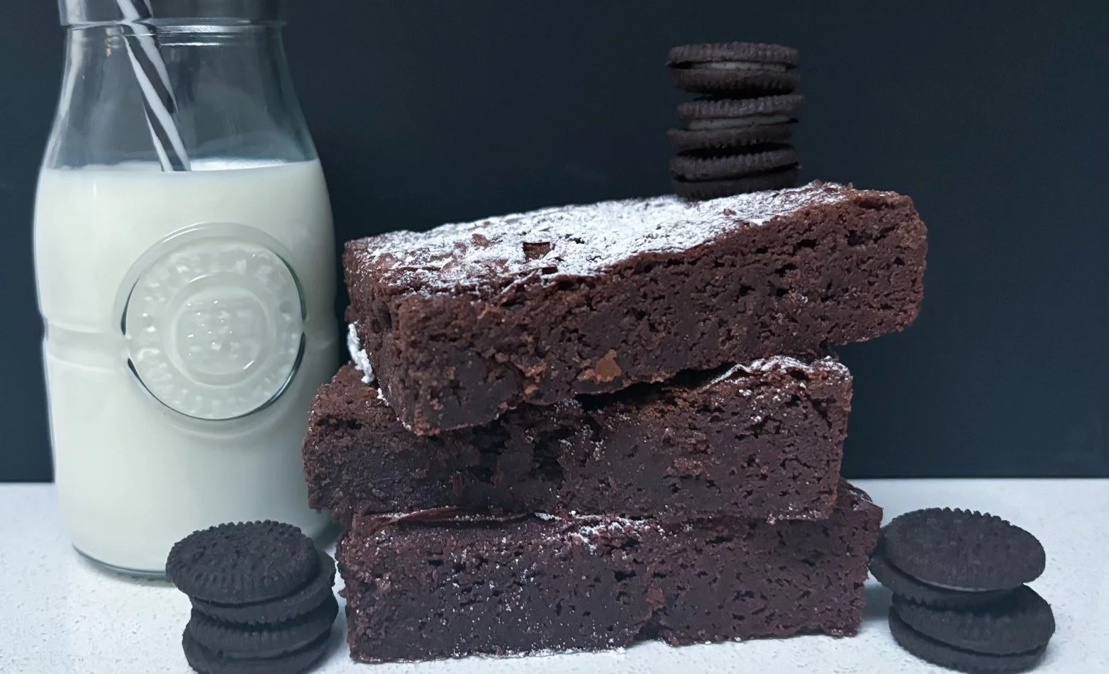 Three stacked chocolate brownies with powdered sugar, surrounded by Oreo cookies on a white surface, with a bottle of milk nearby against a dark background.