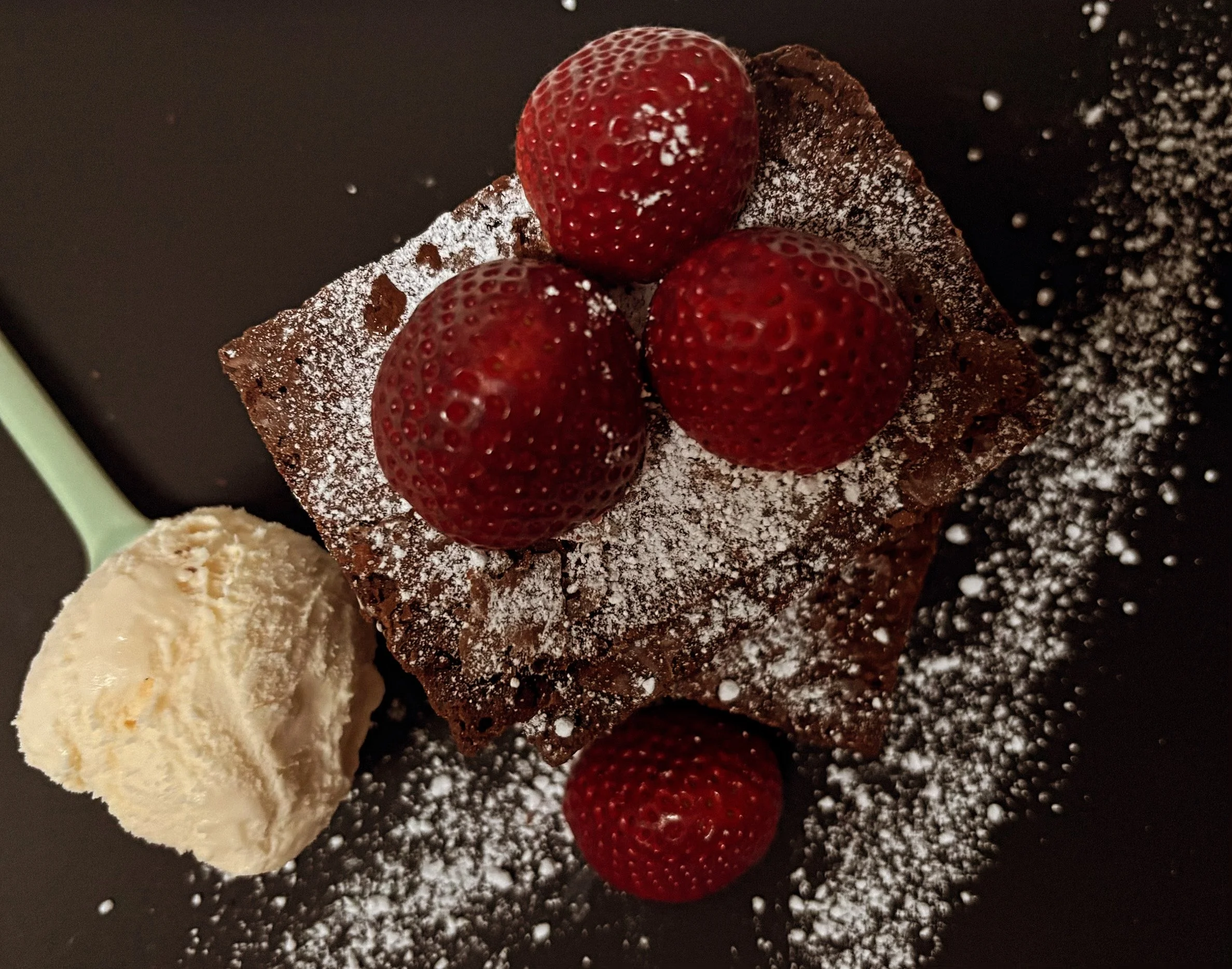 Chocolate brownie topped with three strawberries and powdered sugar, served with a scoop of vanilla ice cream.