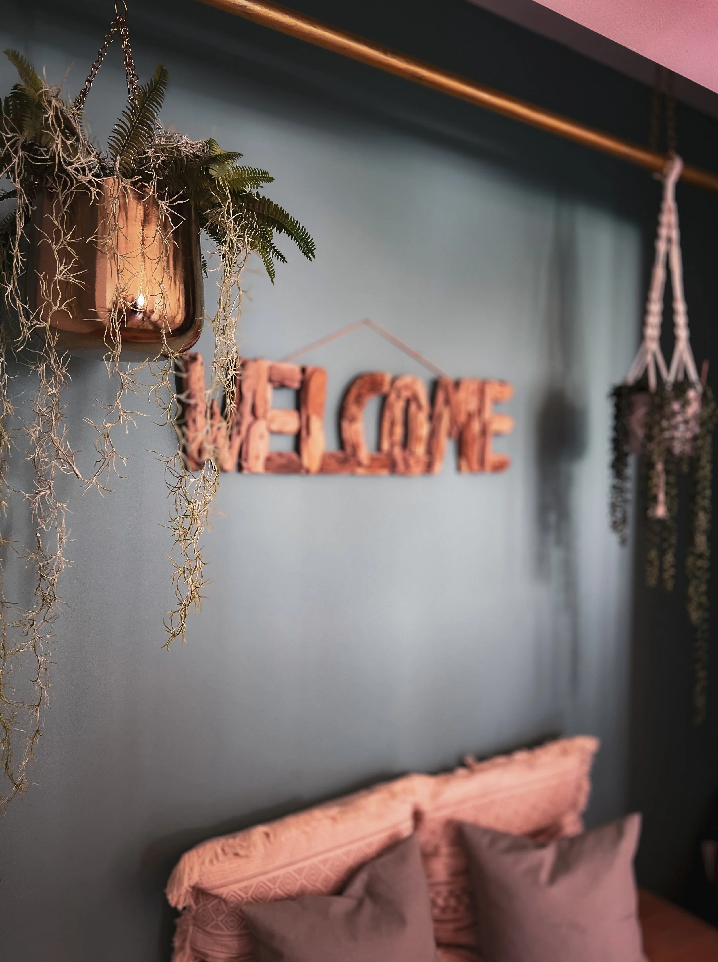 Décoration intérieure avec un mur bleu, comprenant un signe 'HOME' en bois, deux plantes suspendues et des oreillers sur un lit.