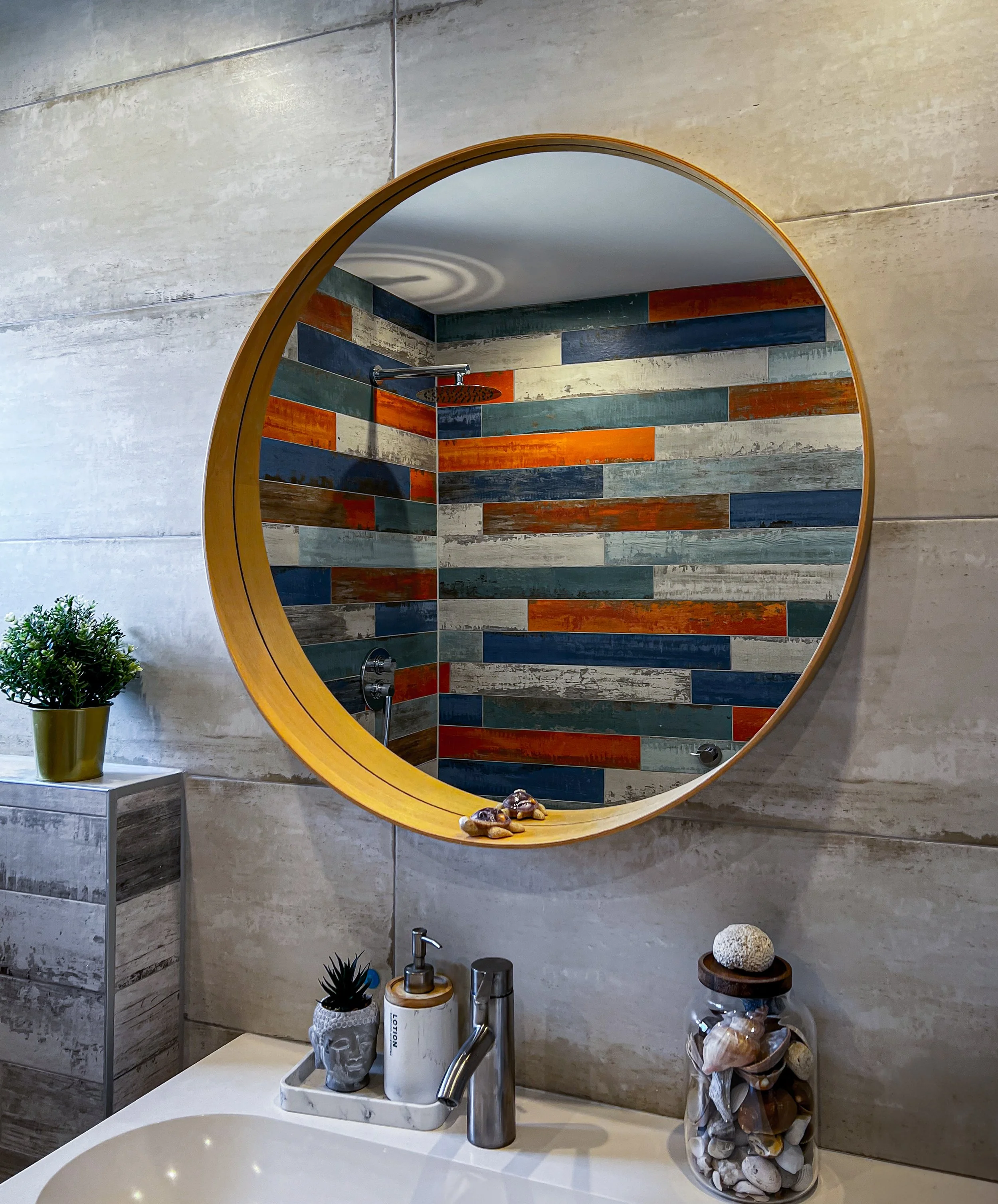 Un miroir rond au-dessus d'un lavabo, avec une décoration de coquillages et de plantes en pot. La pièce reflète une douche avec un mur en carreaux de différentes couleurs, notamment le bleu, l'orange, le gris et le blanc.