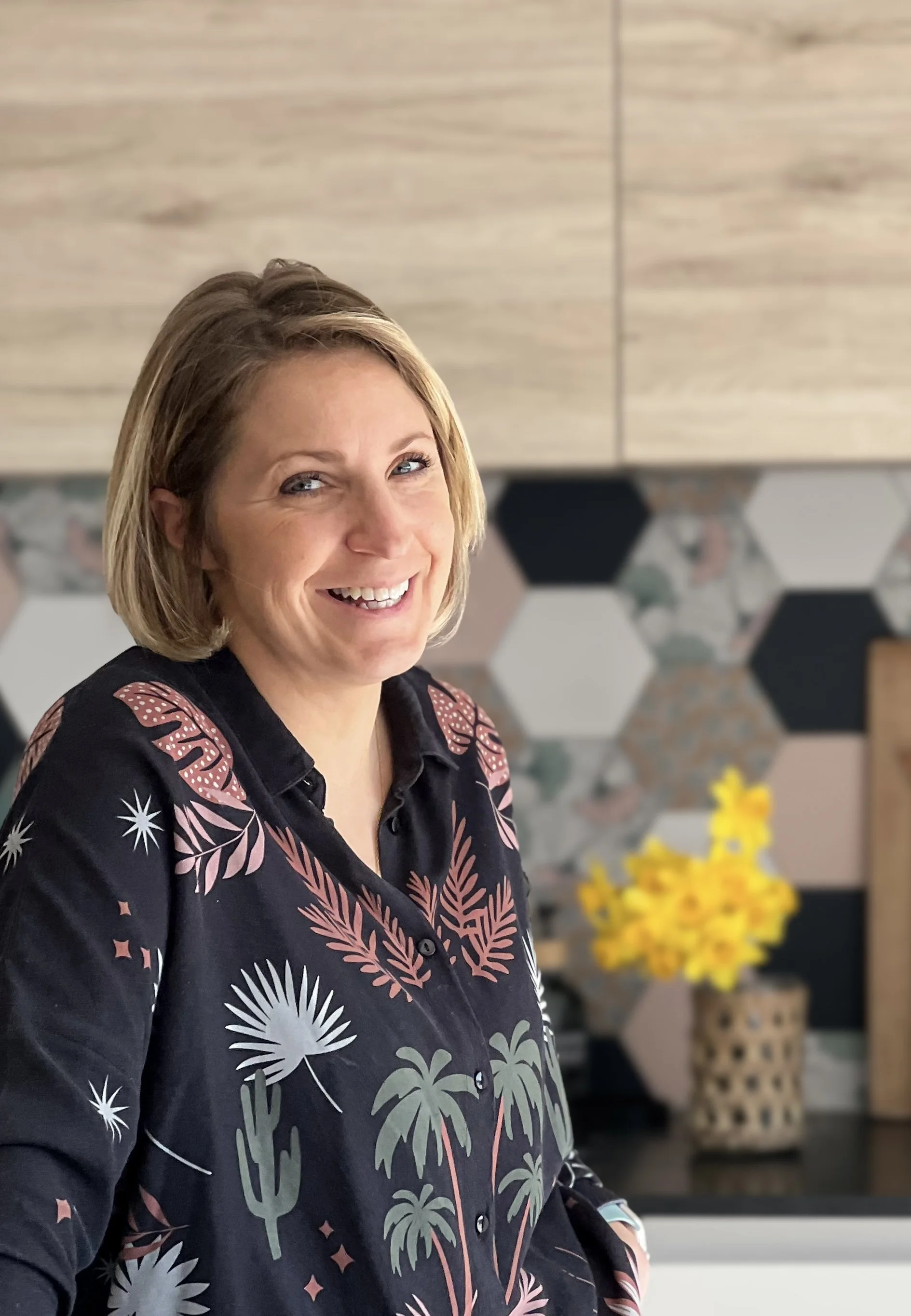 Une femme souriante avec des cheveux courts, portant une chemise noire avec un motif de palmiers et de feuilles, dans une cuisine moderne avec un mur en carreaux hexagonaux et un vase de fleurs jaunes en arrière-plan.