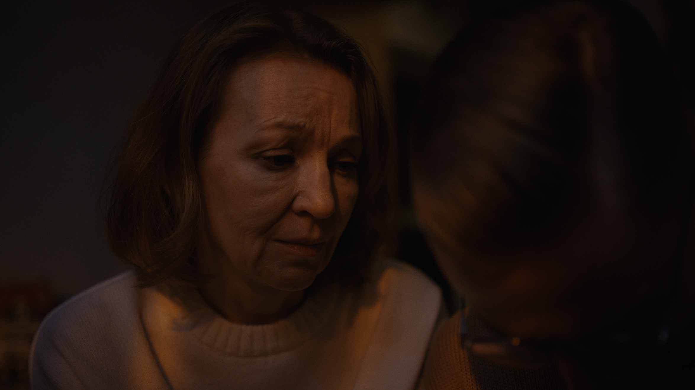 A woman with short, wavy hair looking down, sitting at a table in a dimly lit environment.