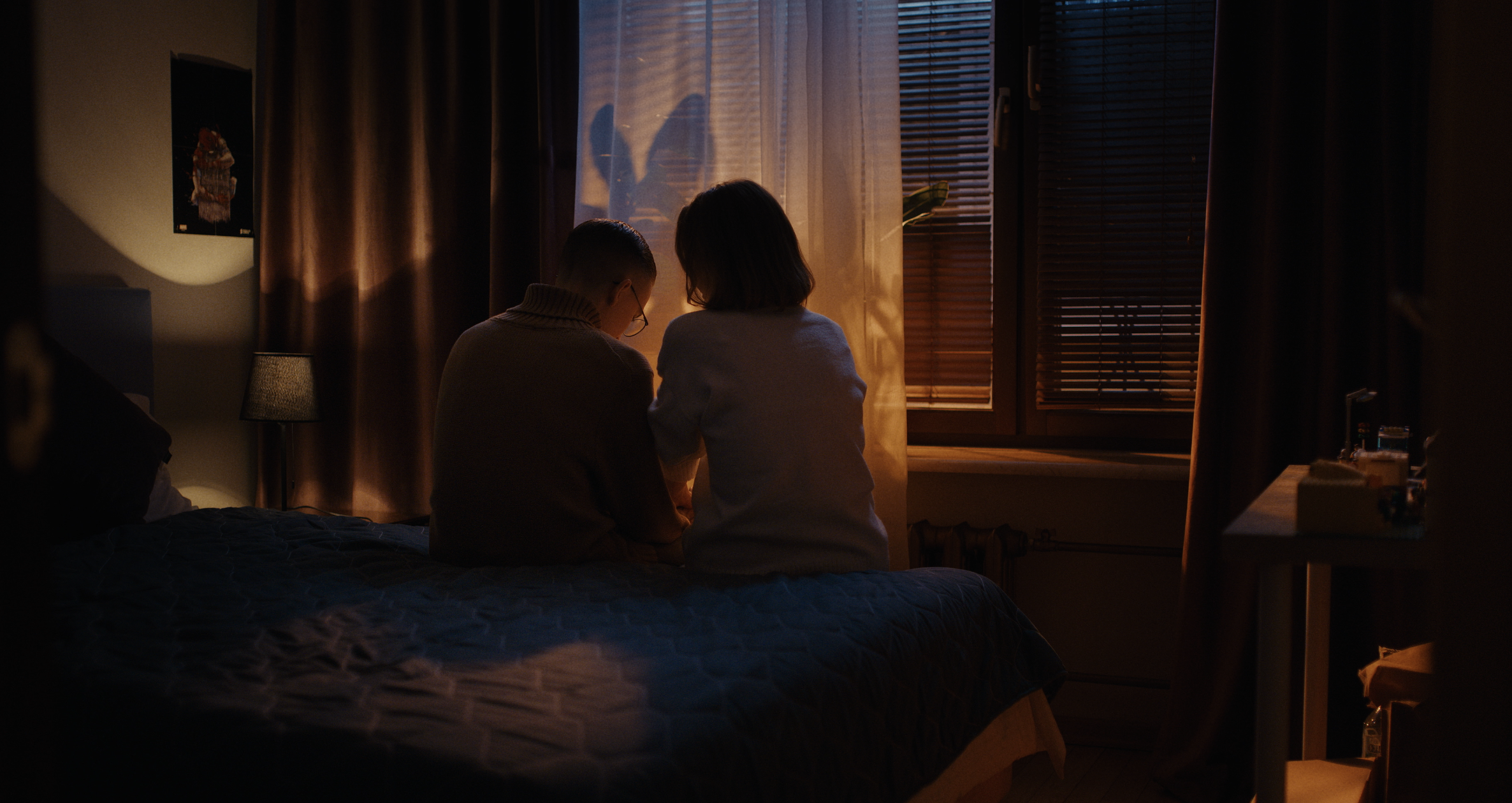 Two people sitting in a bedroom near the window with blinds, backlit by sunset, shadows cast on the wall, and a small lamp on the bedside table.