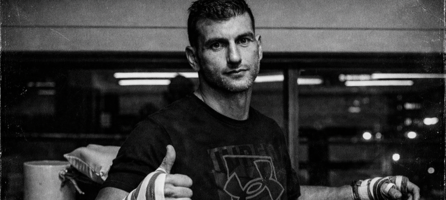 Black and white photo of a man giving a thumbs up, standing in an indoor setting with a casual look, wearing a t-shirt and wristbands.