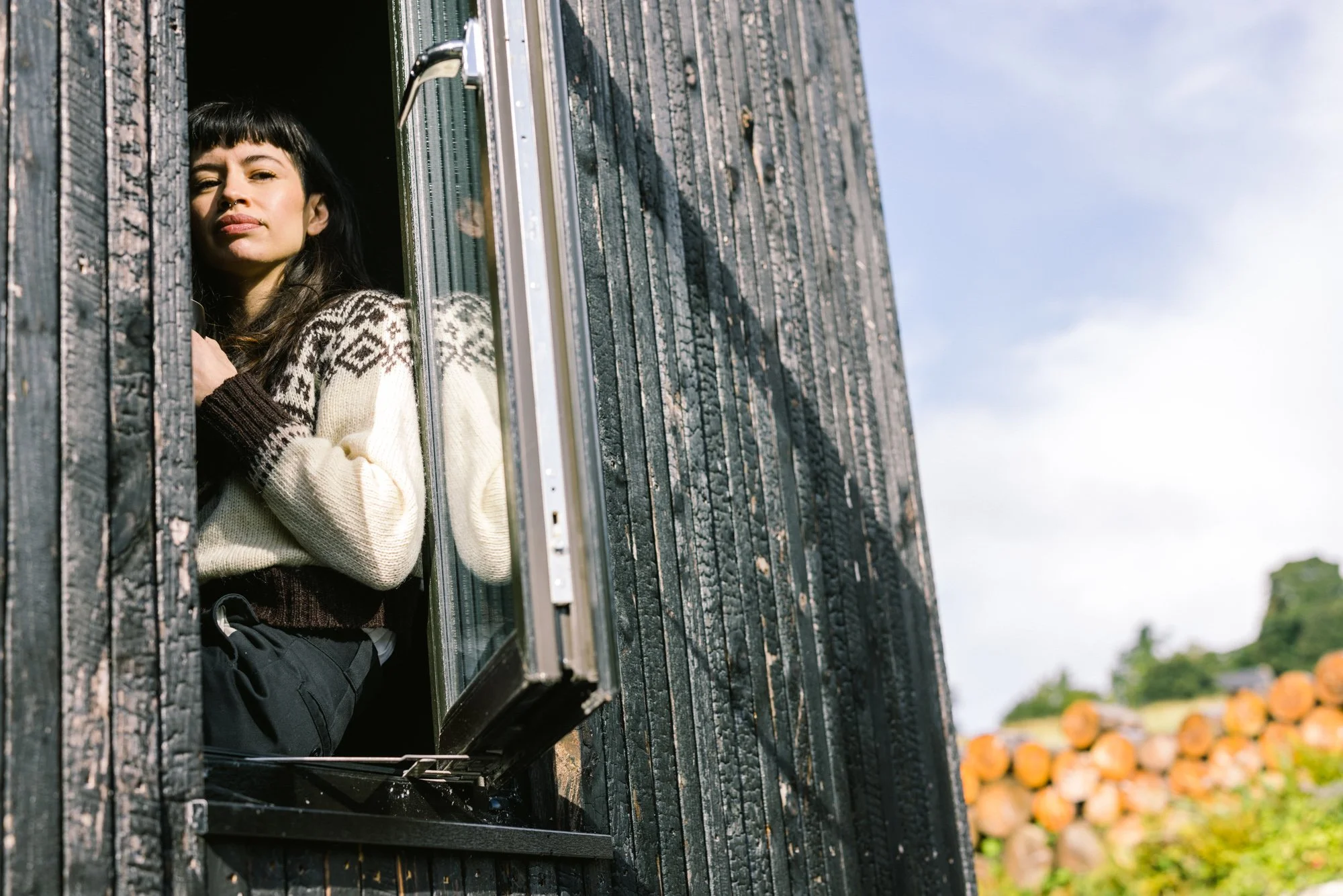 Joven mujer asomándose por la ventana de una casa de madera con cielo despejado en el fondo.