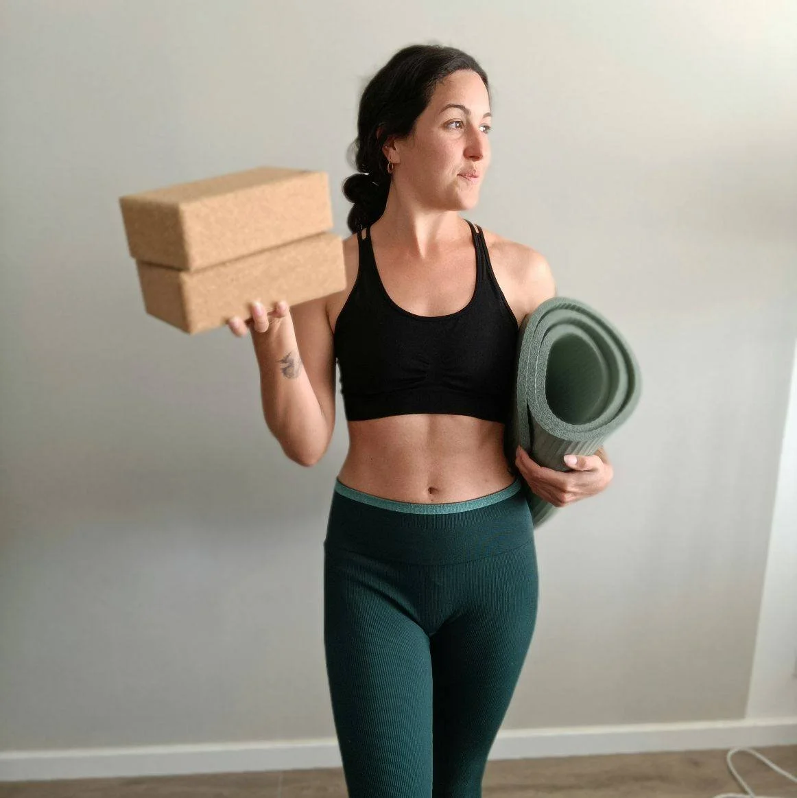 Femme sportive tenant deux briques dans une main et un tapis de yoga dans l'autre, portant une tenue de sport noire et vert foncé, dans une pièce neutre.