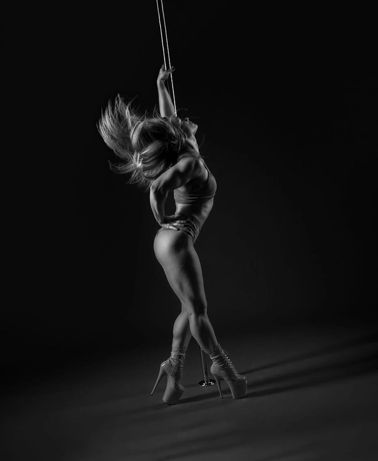 A woman with long blonde hair performing a pole dance move on a hoop outdoors under a patio roof, with colorful flowers and greenery in the background.