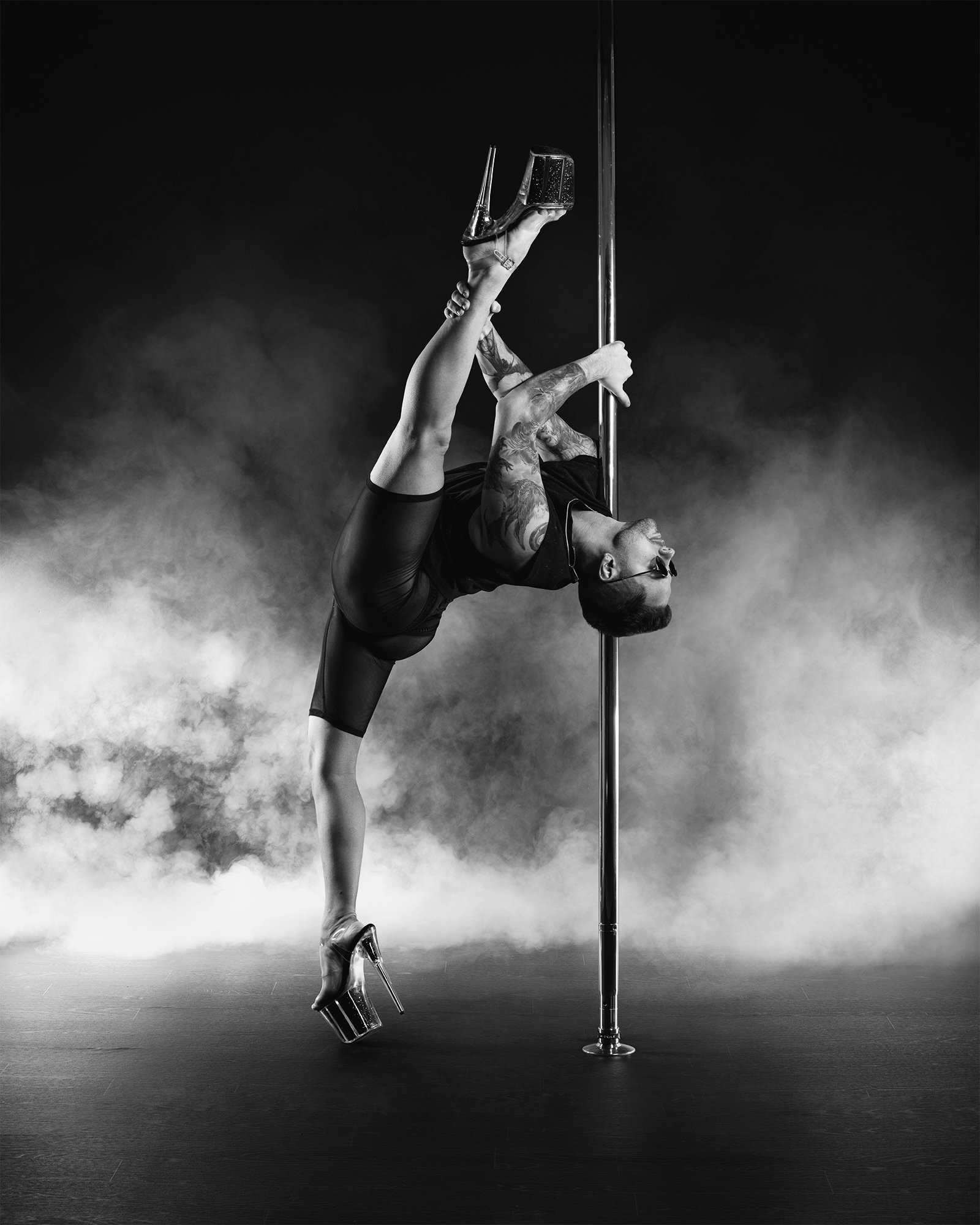 A woman performing a pole dance move, hanging upside down with her legs draped around the pole, wearing a gold-colored outfit against a plain background.