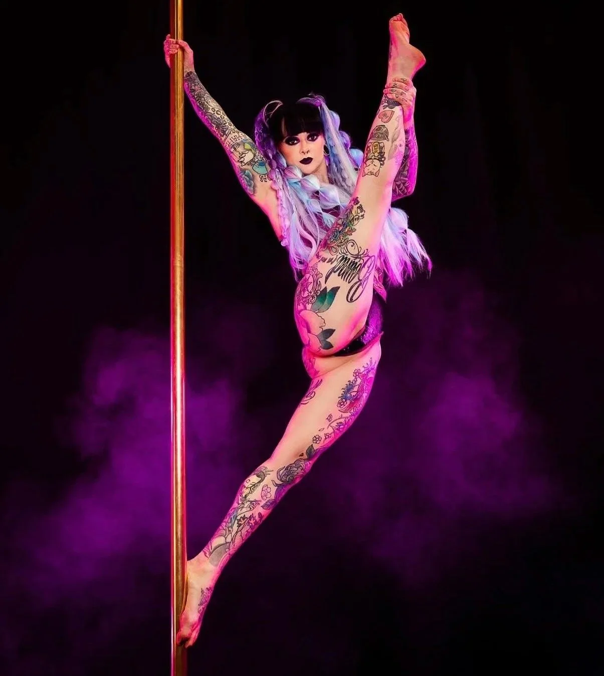 A woman performing a pole dance move, hanging upside down with her legs draped around the pole, wearing a gold-colored outfit against a plain background.
