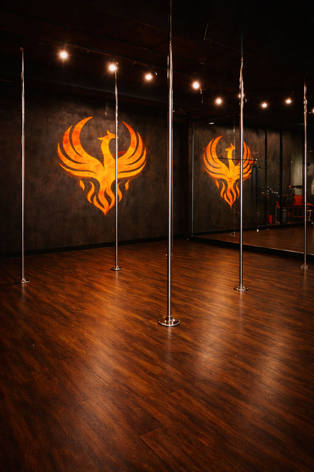 Dance studio with wooden floor, black wall with orange phoenix logo, and mirror reflecting the logo and poles.