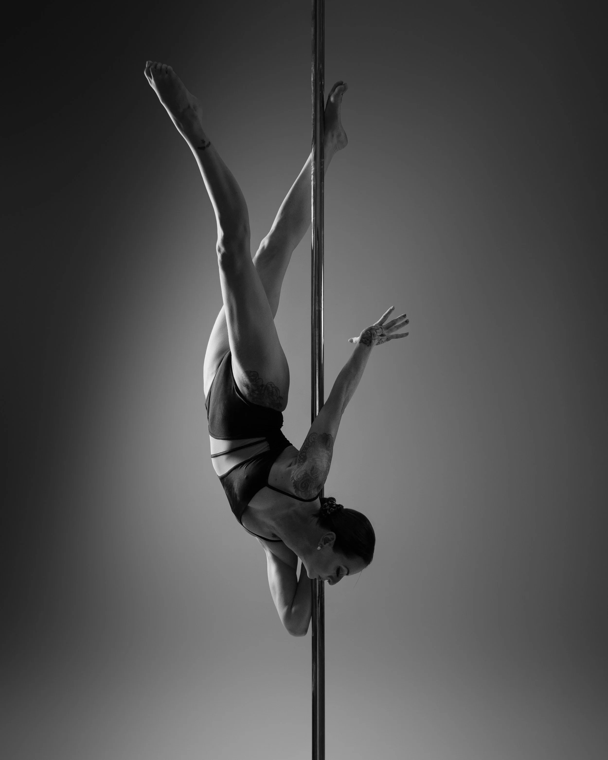 A woman performing a pole dance move on a vertical pole, holding her legs with her hands, in a dimly lit room with a red fabric hanging behind her.
