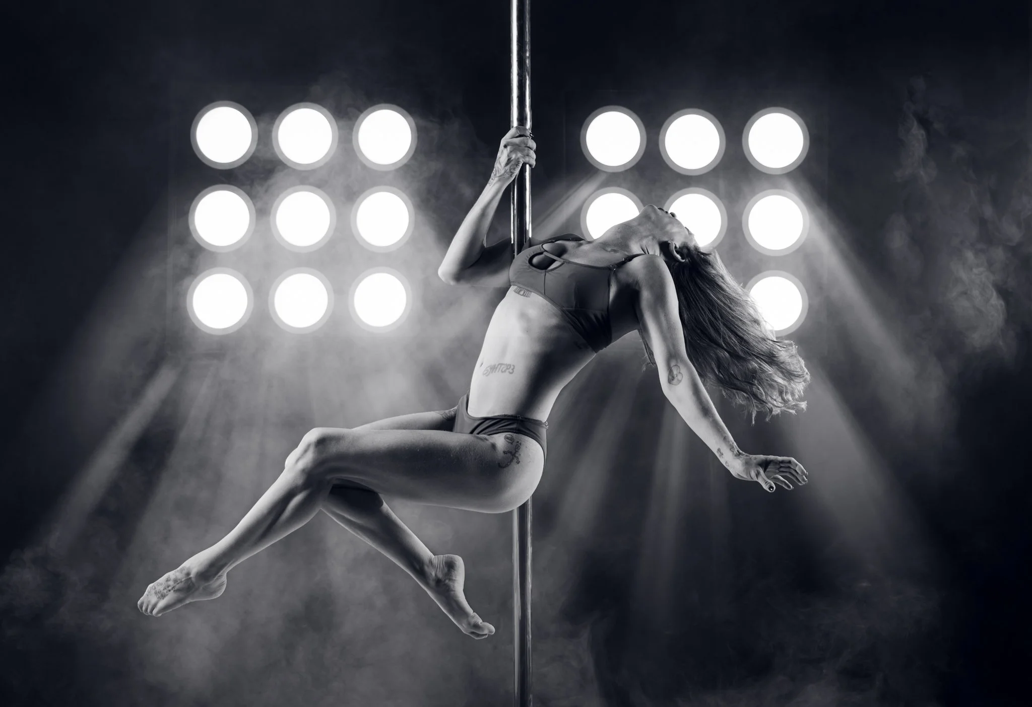 A female pole dancer performing an aerial move on a pole, wearing a white and black costume, against a dark background.